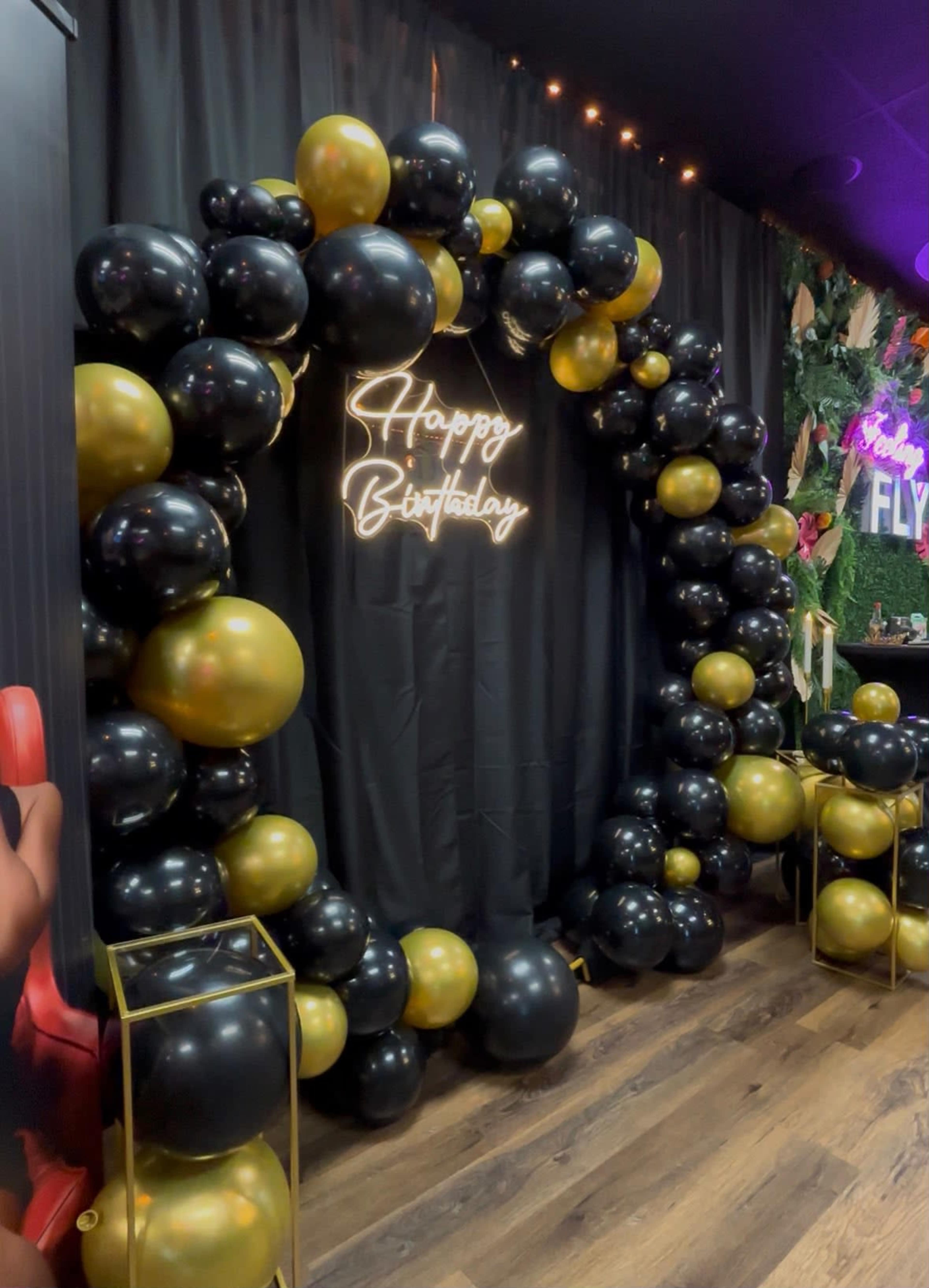 The image features a birthday decoration made of black and gold balloons arranged in a heart shape, with a neon sign that says "Happy Birthday" in the center.