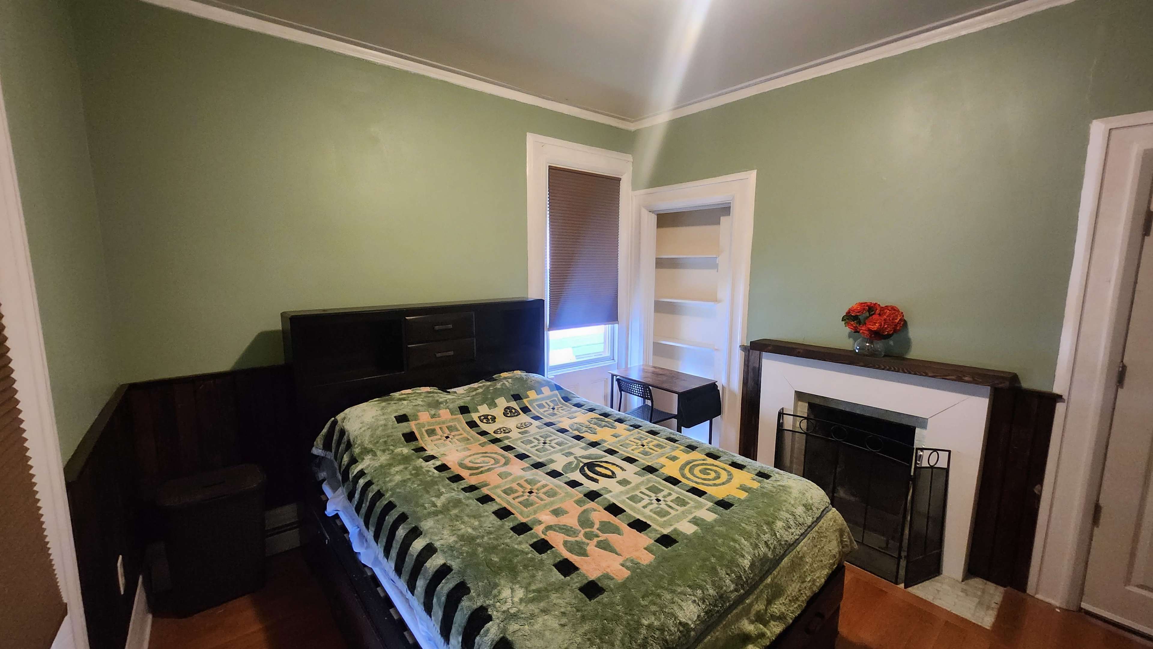 A bedroom featuring a bed with a green and patterned blanket, a small table, and a fireplace with decorative flowers above it.