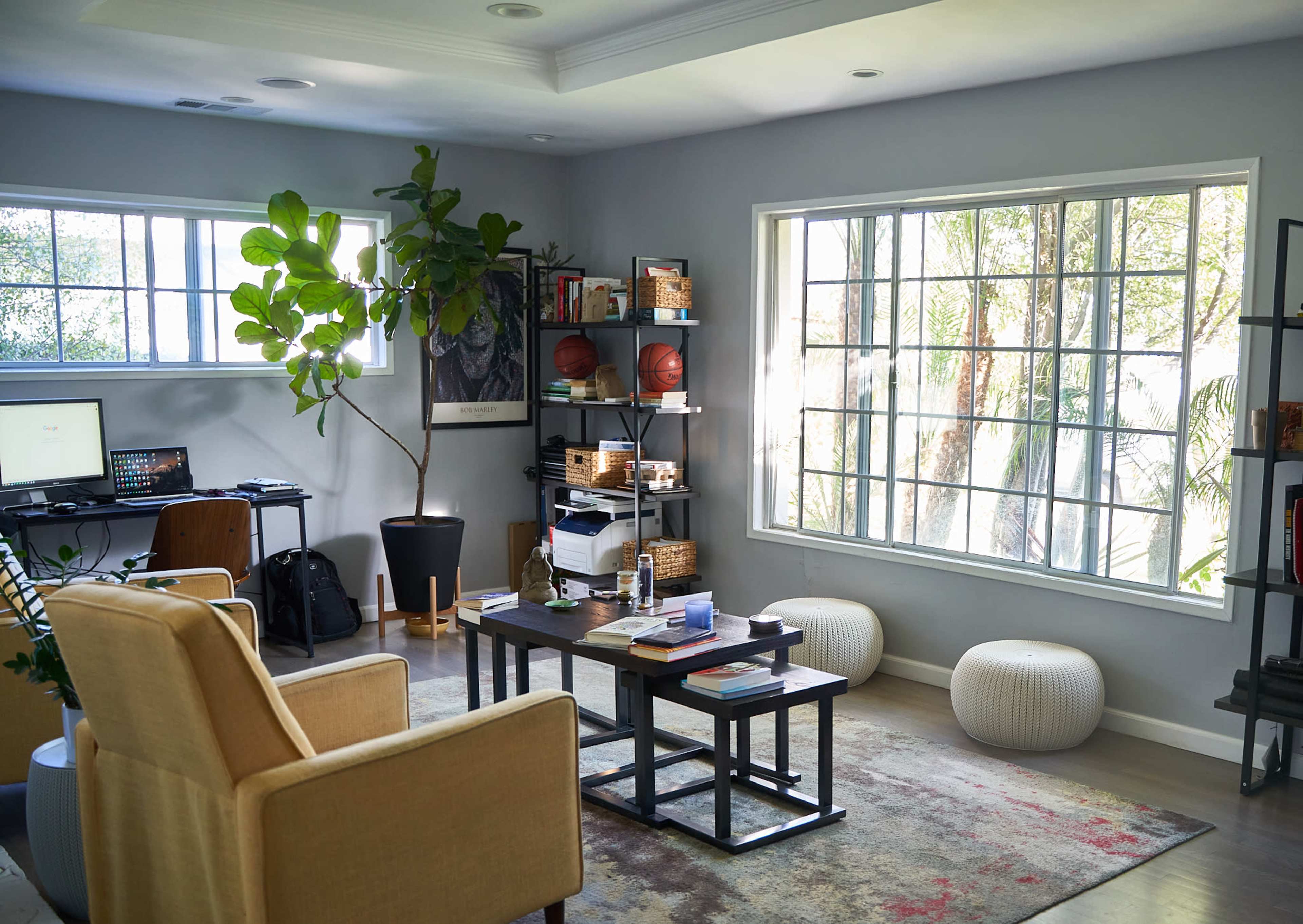 A modern living space features a desk with a computer, shelves filled with books and sports equipment, and large windows letting in natural light.
