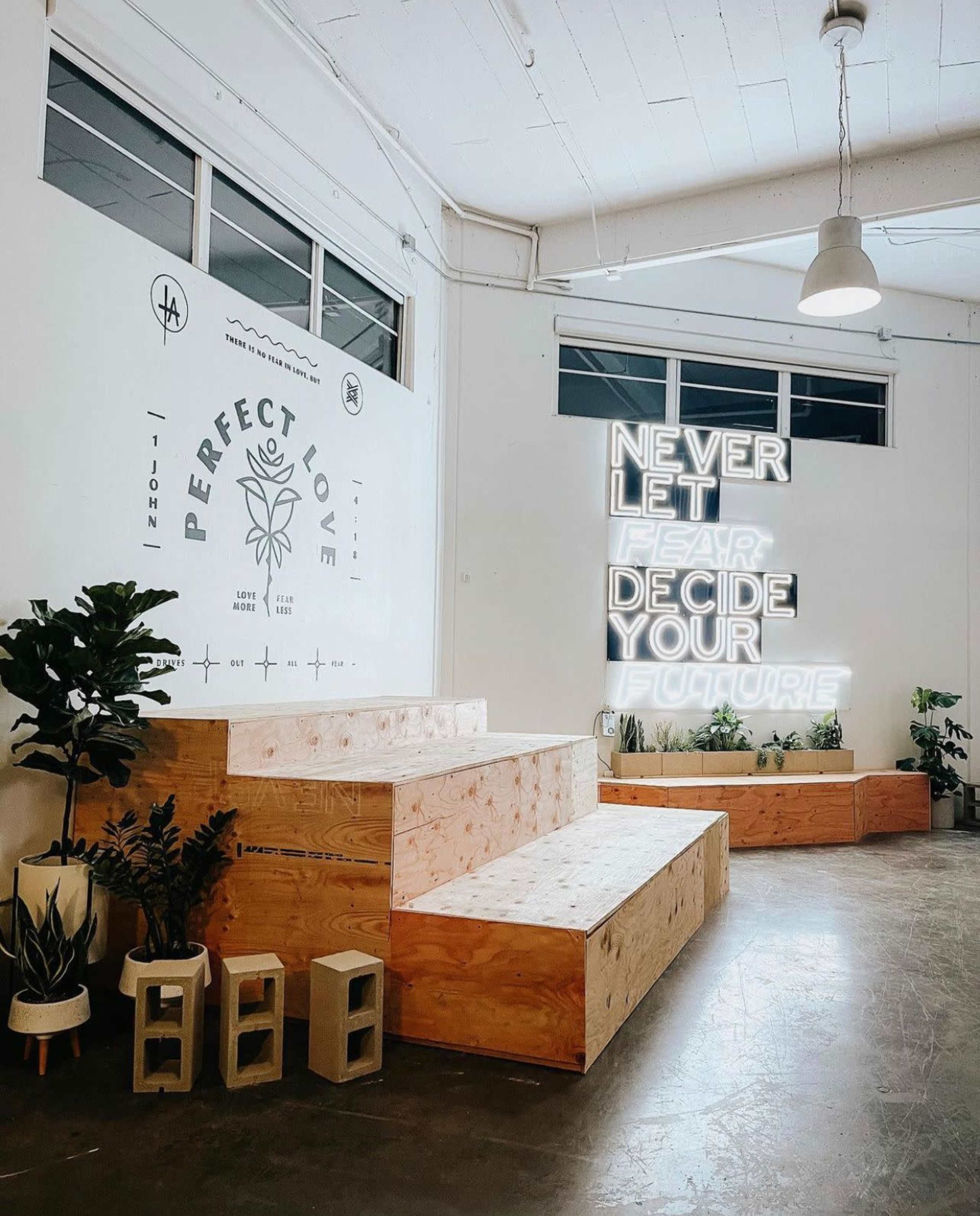 A minimalist interior space featuring wooden seating, plants, and a neon sign that reads "NEVER LET FEAR DECIDE YOUR FUTURE."