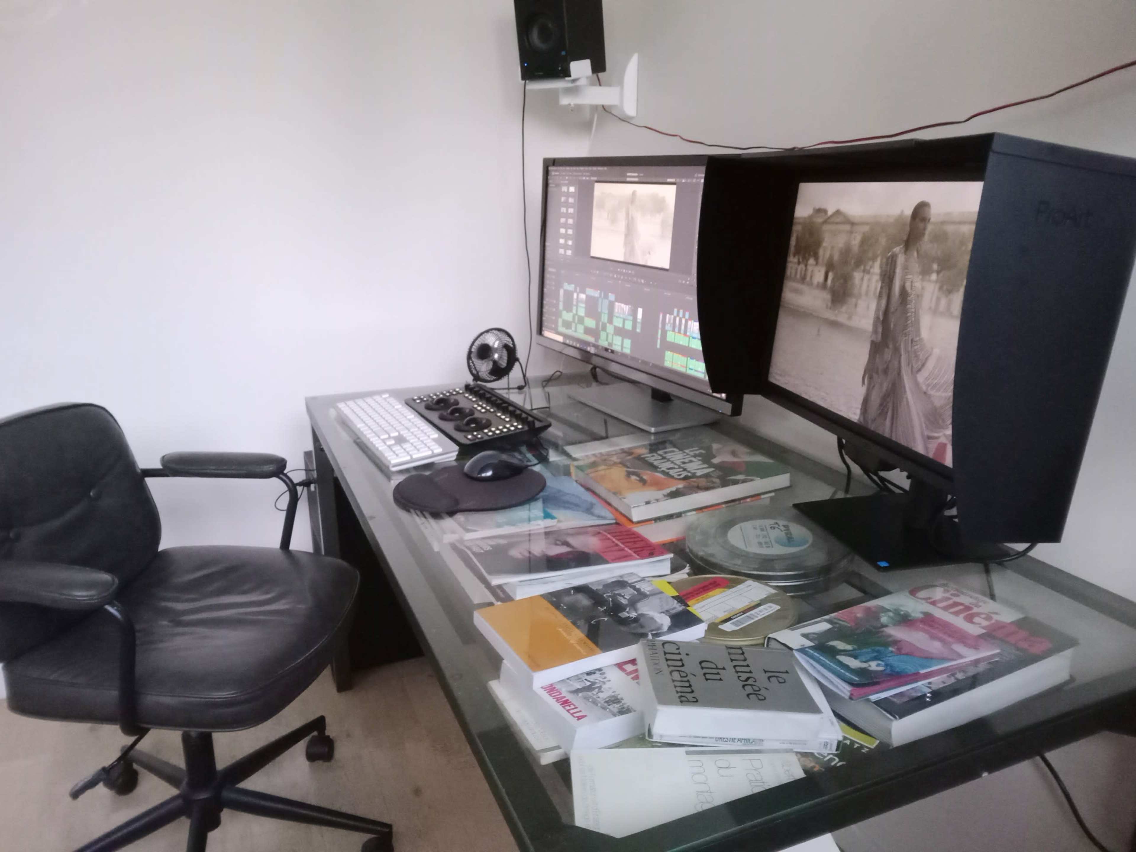 A workstation featuring two large computer monitors, a keyboard, and a stack of books and magazines on a glass tabletop, with a black office chair positioned nearby.