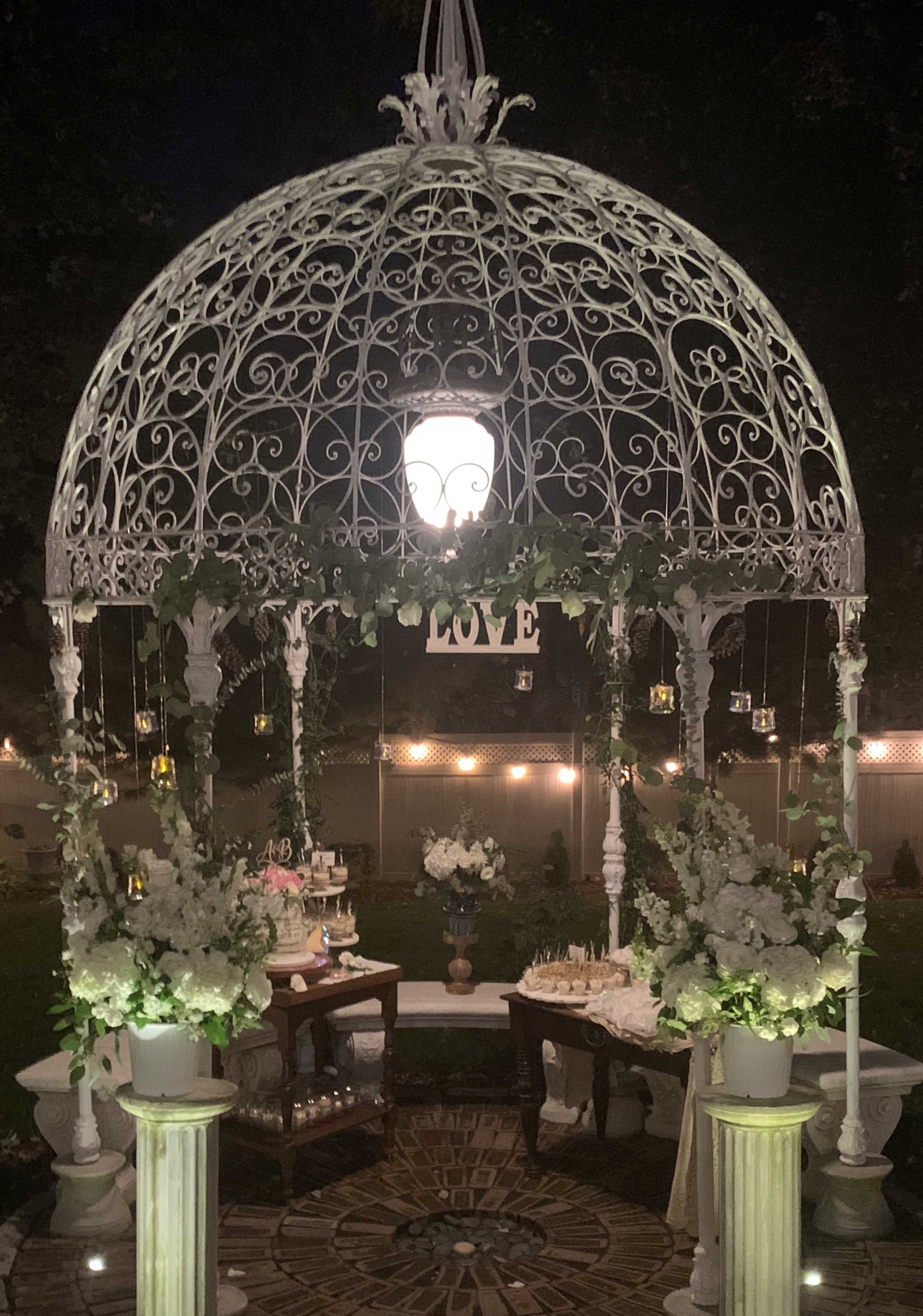 A white gazebo adorned with greenery and flowers is set up for an event, illuminated by soft lights and featuring tables with decorative items.