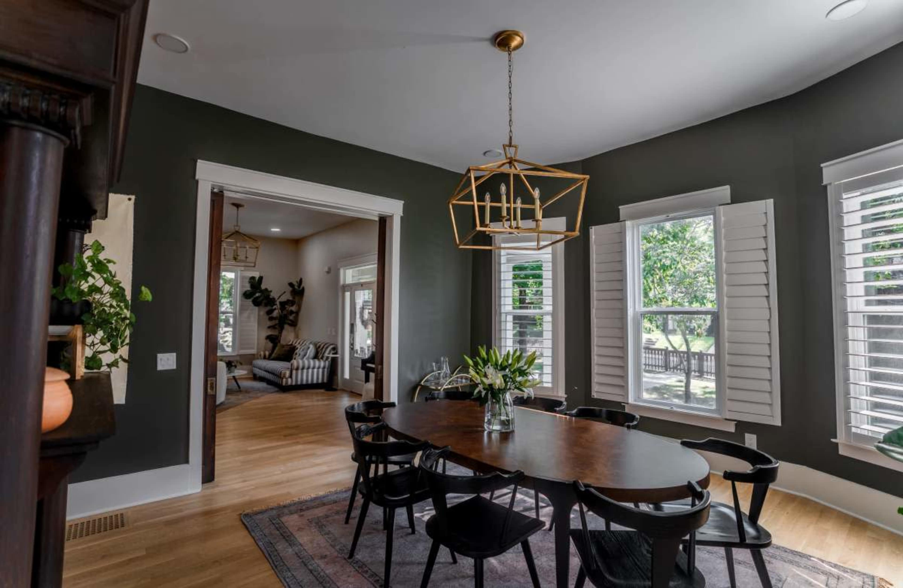 The image shows a dining area with a large table surrounded by black chairs, featuring a modern light fixture and large windows with shutters, leading to a connected living space.