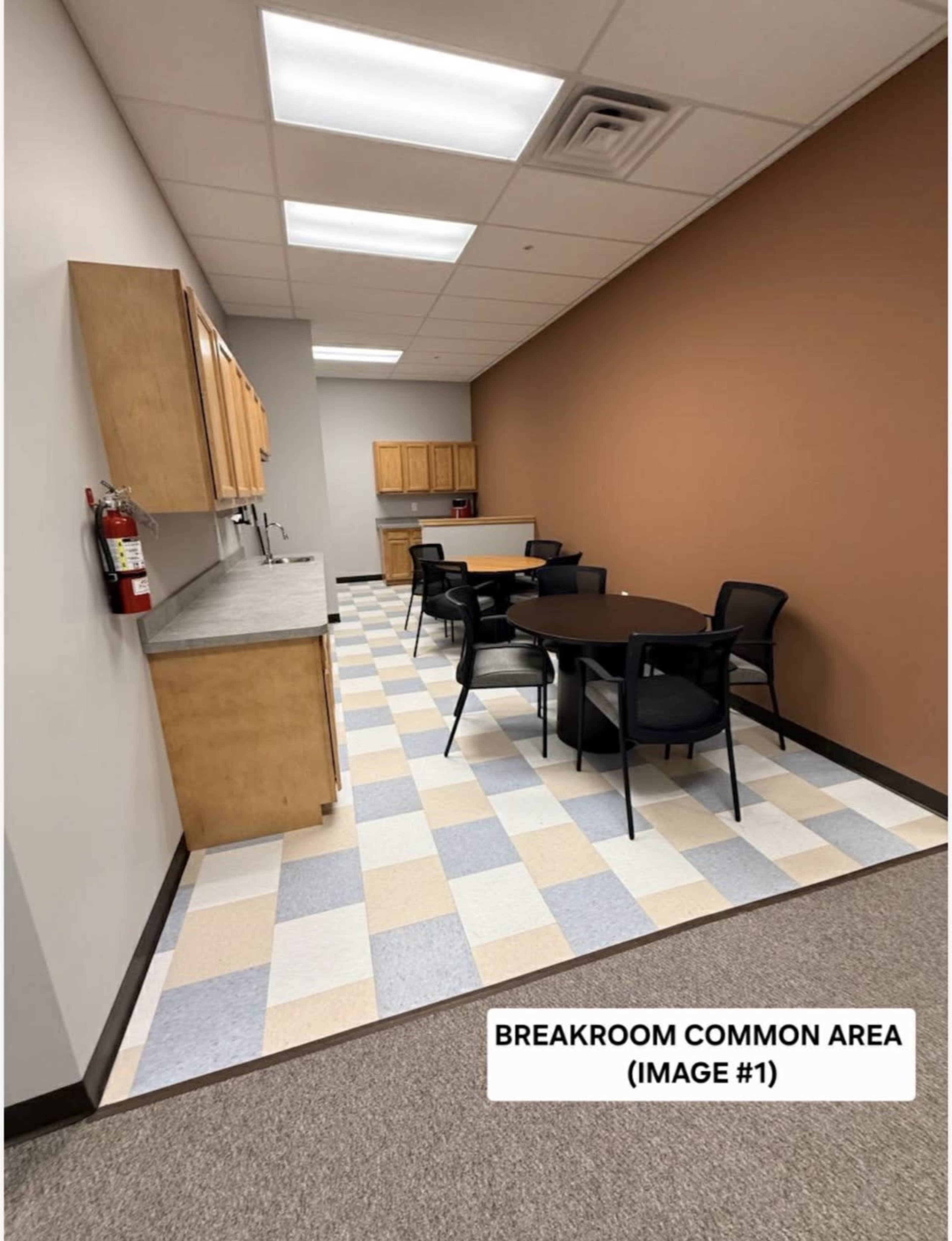 The image shows a breakroom common area with a light-colored tiled floor, a kitchen counter with cabinets, and several black chairs around circular tables.