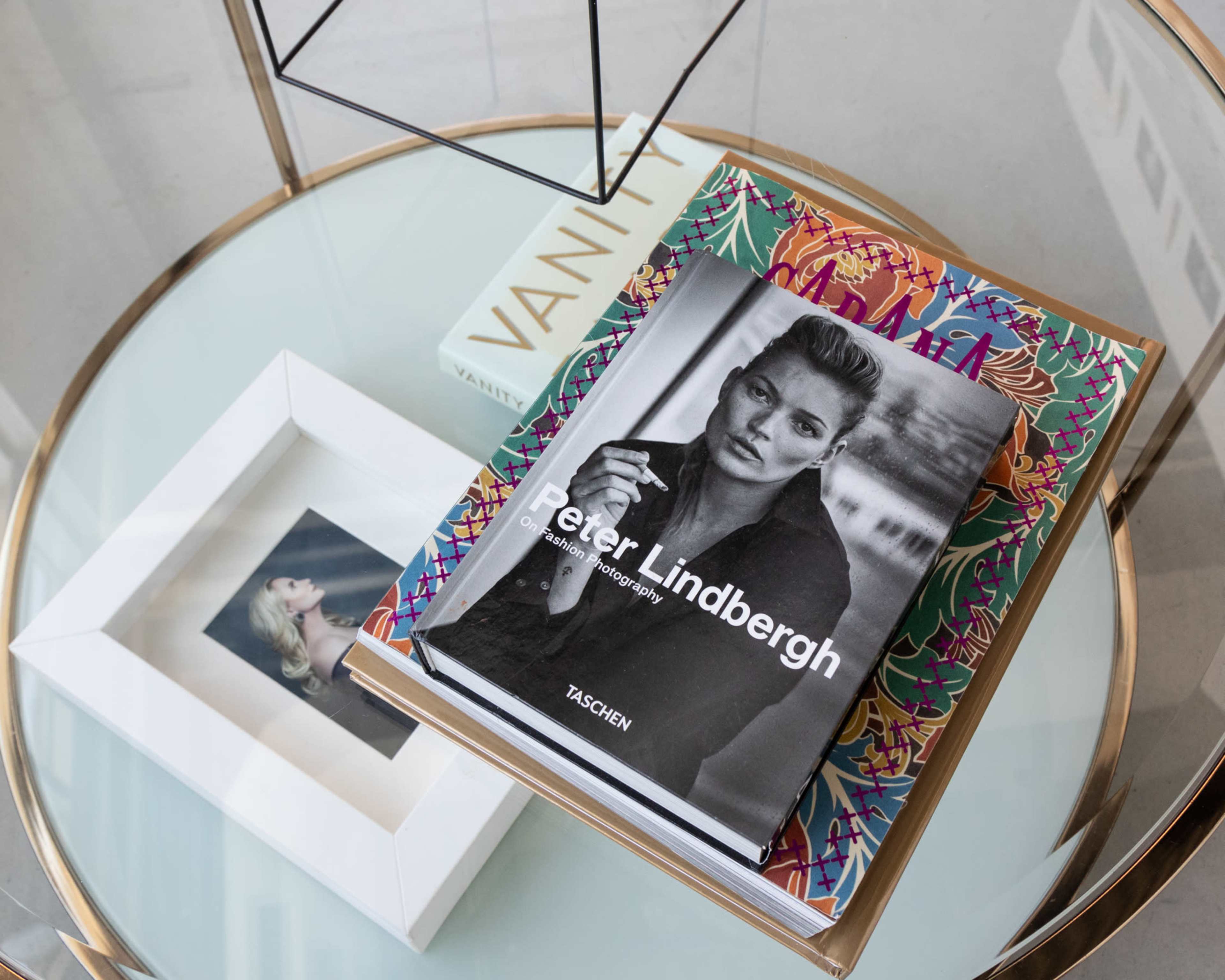 A glass table displays several books, including a photograph book by Peter Lindbergh, alongside a framed picture and a white box labeled "Vanity."