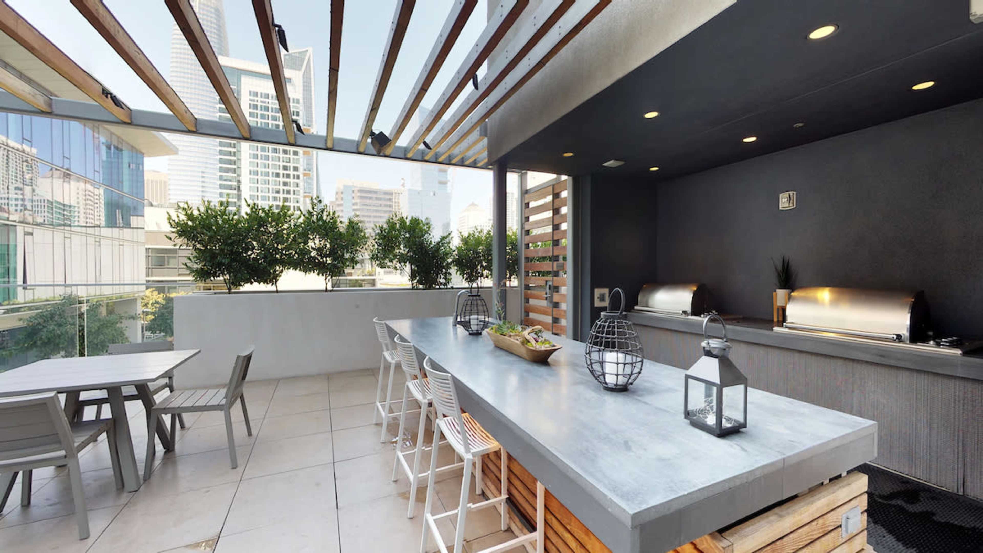 A modern outdoor kitchen area with a long concrete countertop, bar stools, cooking grills, and a dining table beneath a slatted roof.