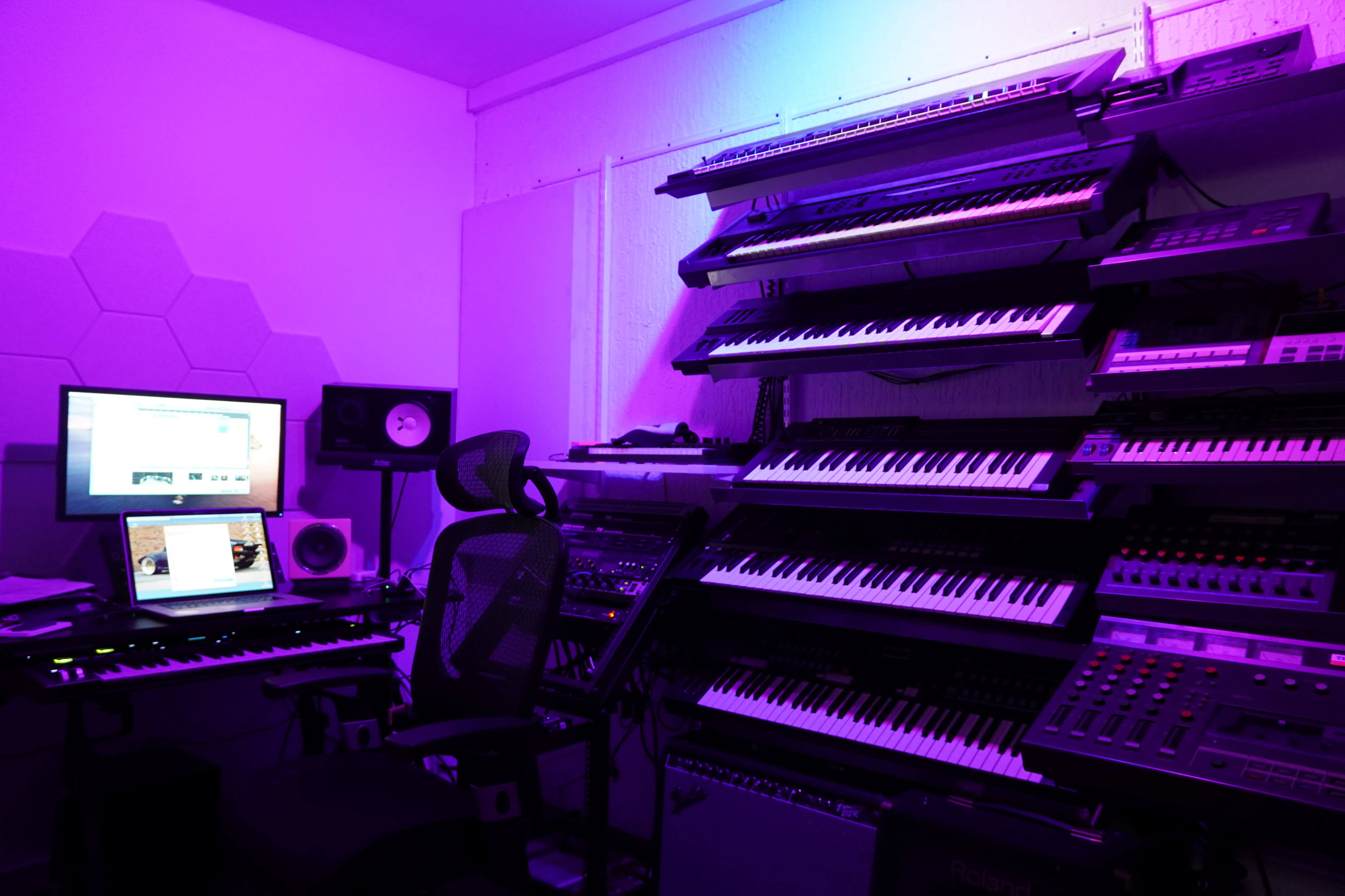 A music production studio features multiple keyboards arranged on shelves with a computer and audio equipment.