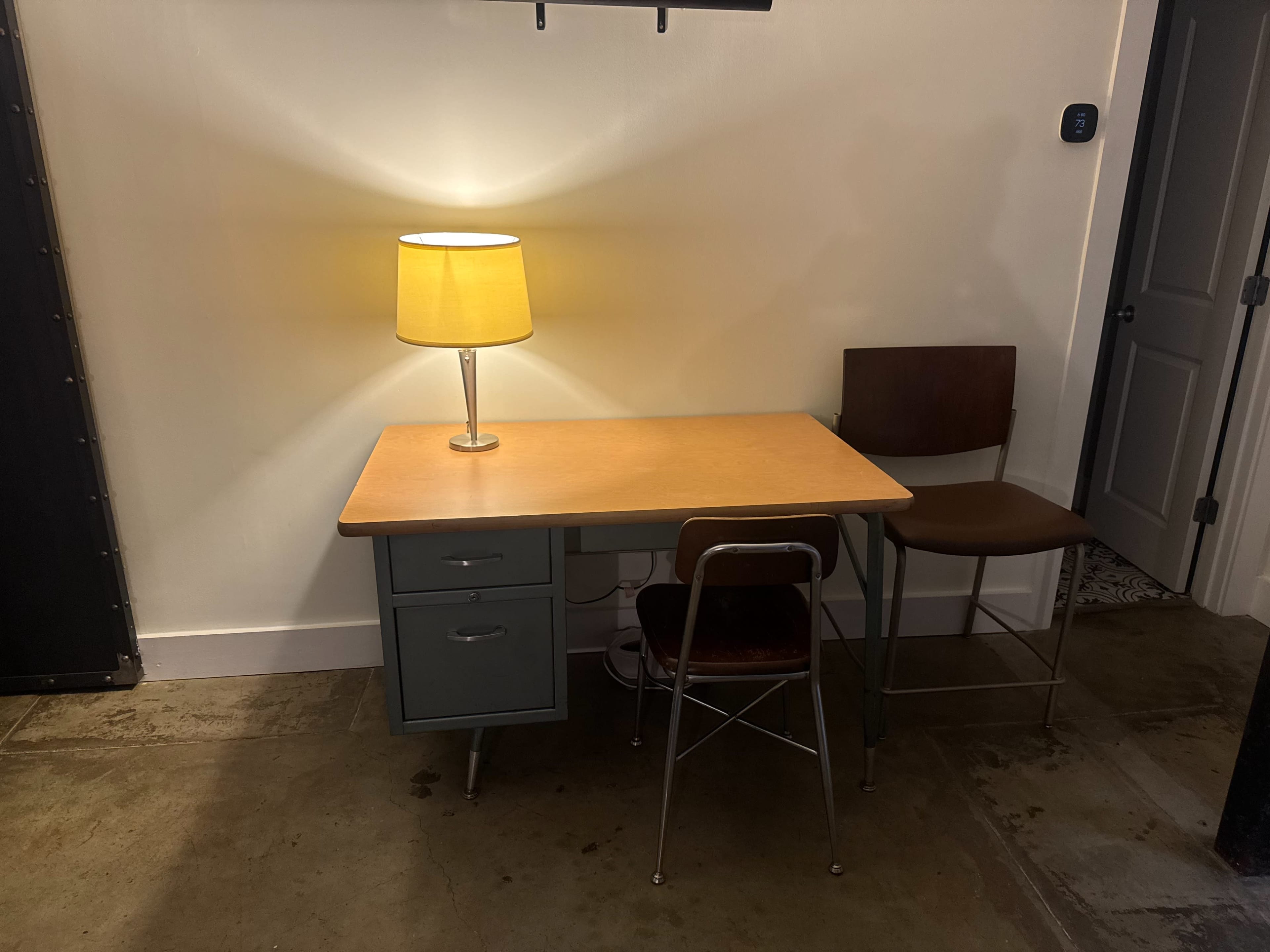 A desk with a wooden top and two drawers is positioned beside a chair and illuminated by a yellow lampshade.