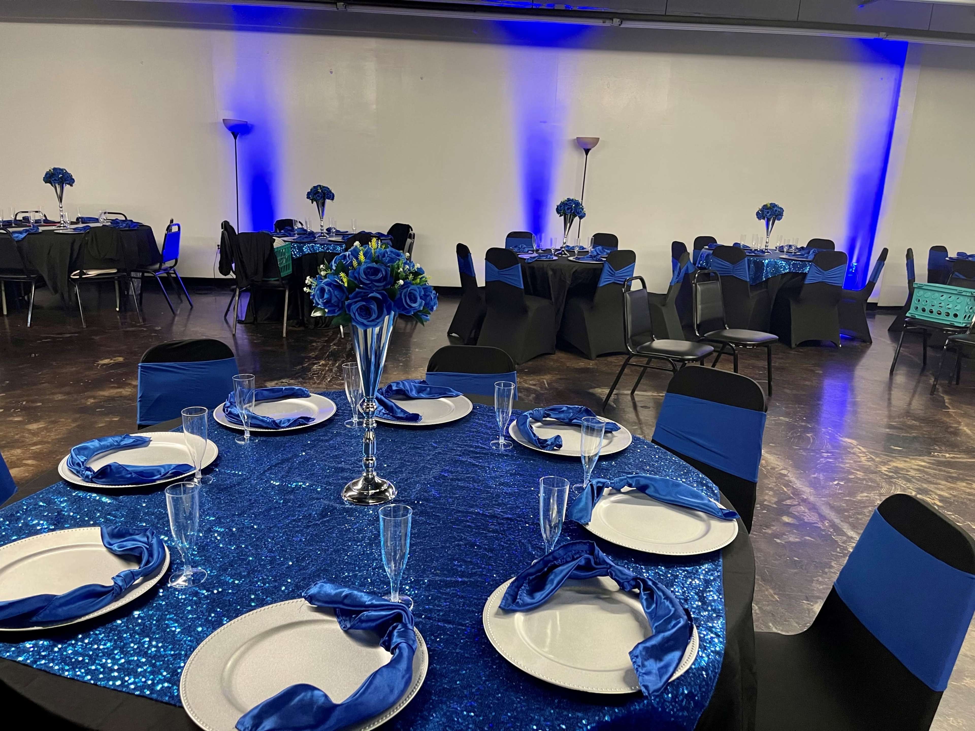 A decorated event space with several tables set with blue and silver decor, including flower centerpieces and silver plates.
