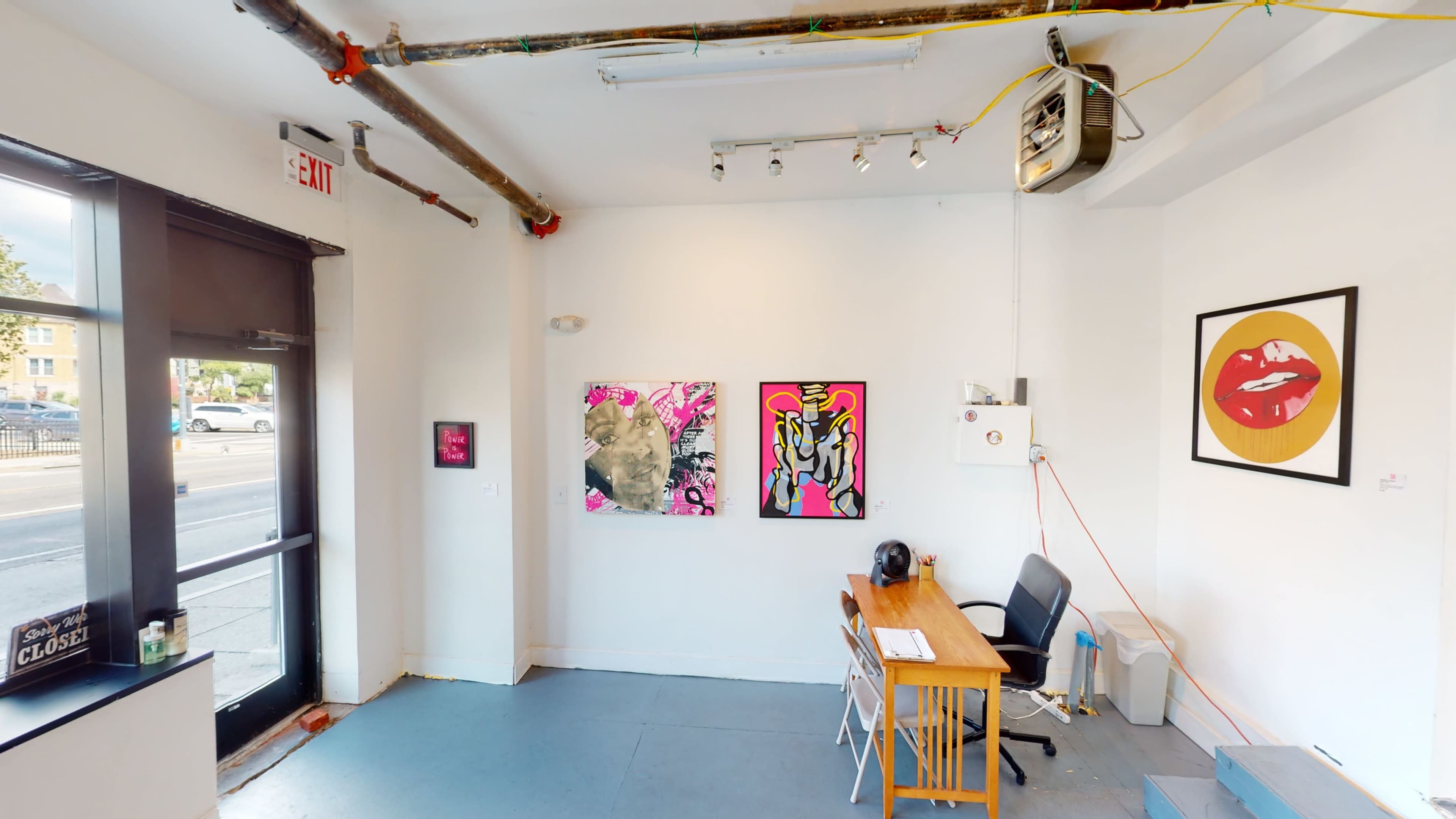 A minimalist art gallery with a wooden table and chair, featuring colorful paintings on the walls and visible street outside through a large window.
