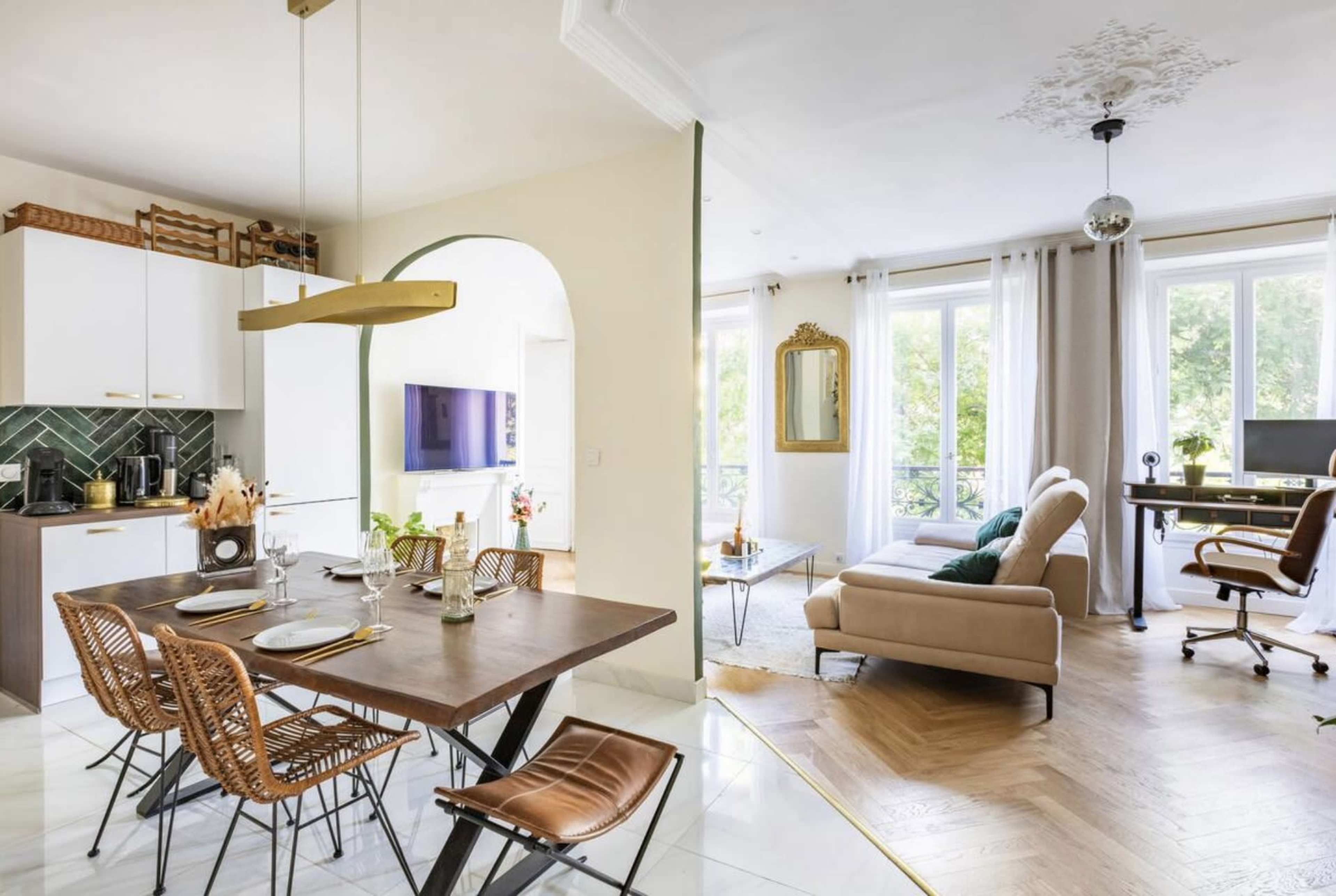 A bright, modern apartment features a dining area with a wooden table and woven chairs, adjacent to a living space with a beige sofa and a workstation near large windows.