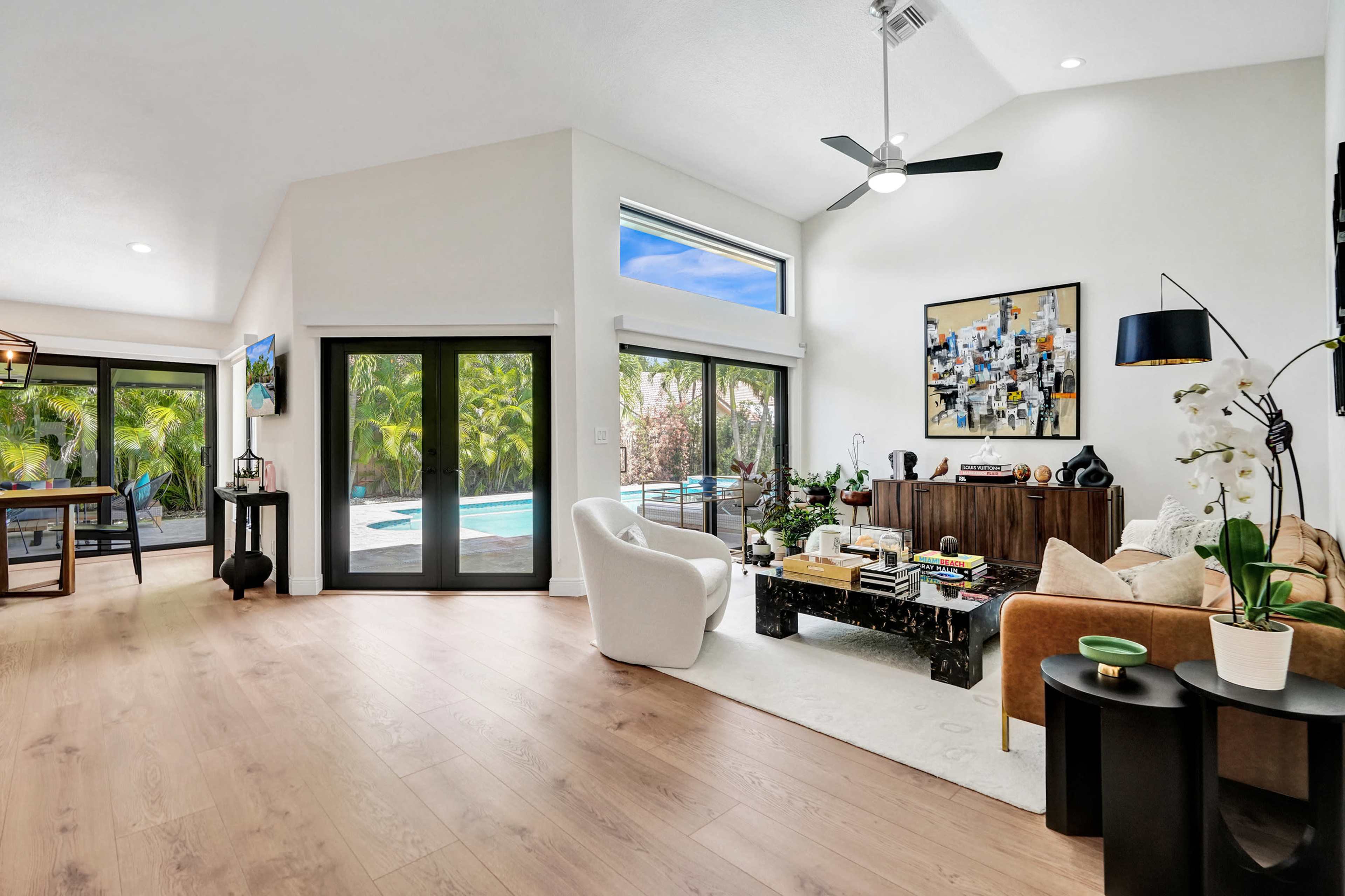 The bright living room features a modern design with large windows showcasing a view of the pool and outdoor greenery.