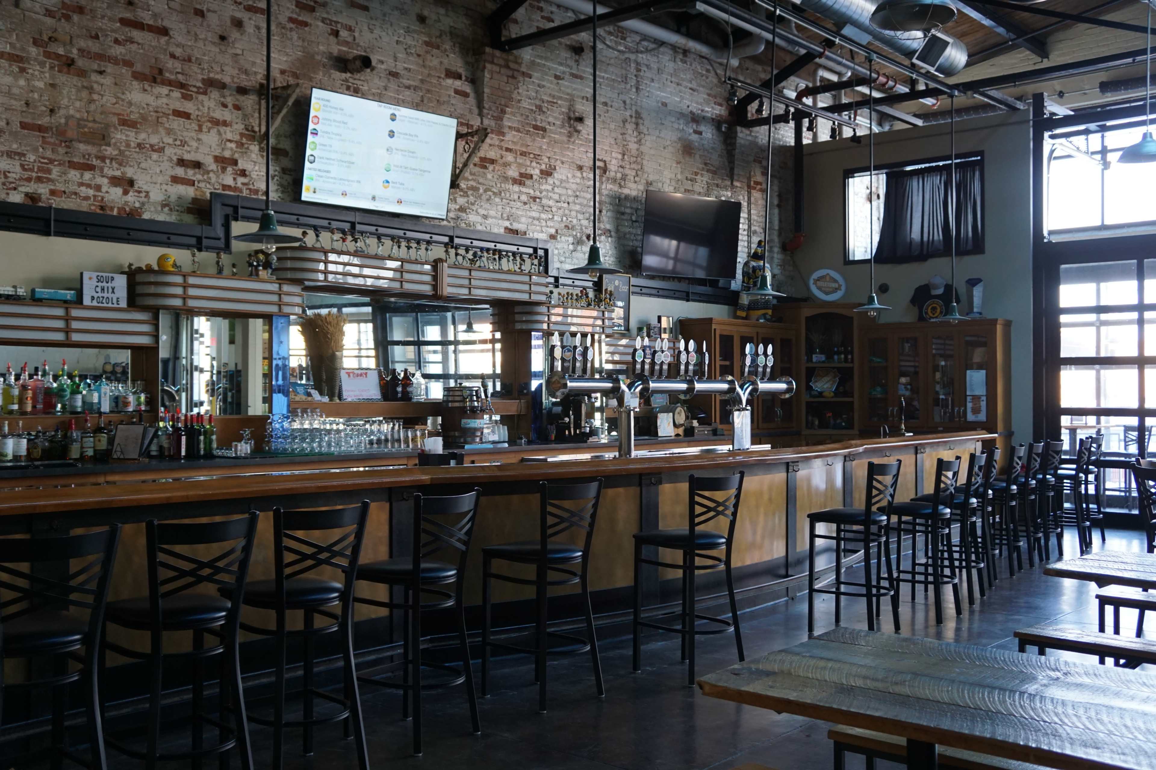 A bar area with a long wooden counter, several high stools, and shelves displaying liquor bottles against a backdrop of exposed brick walls.