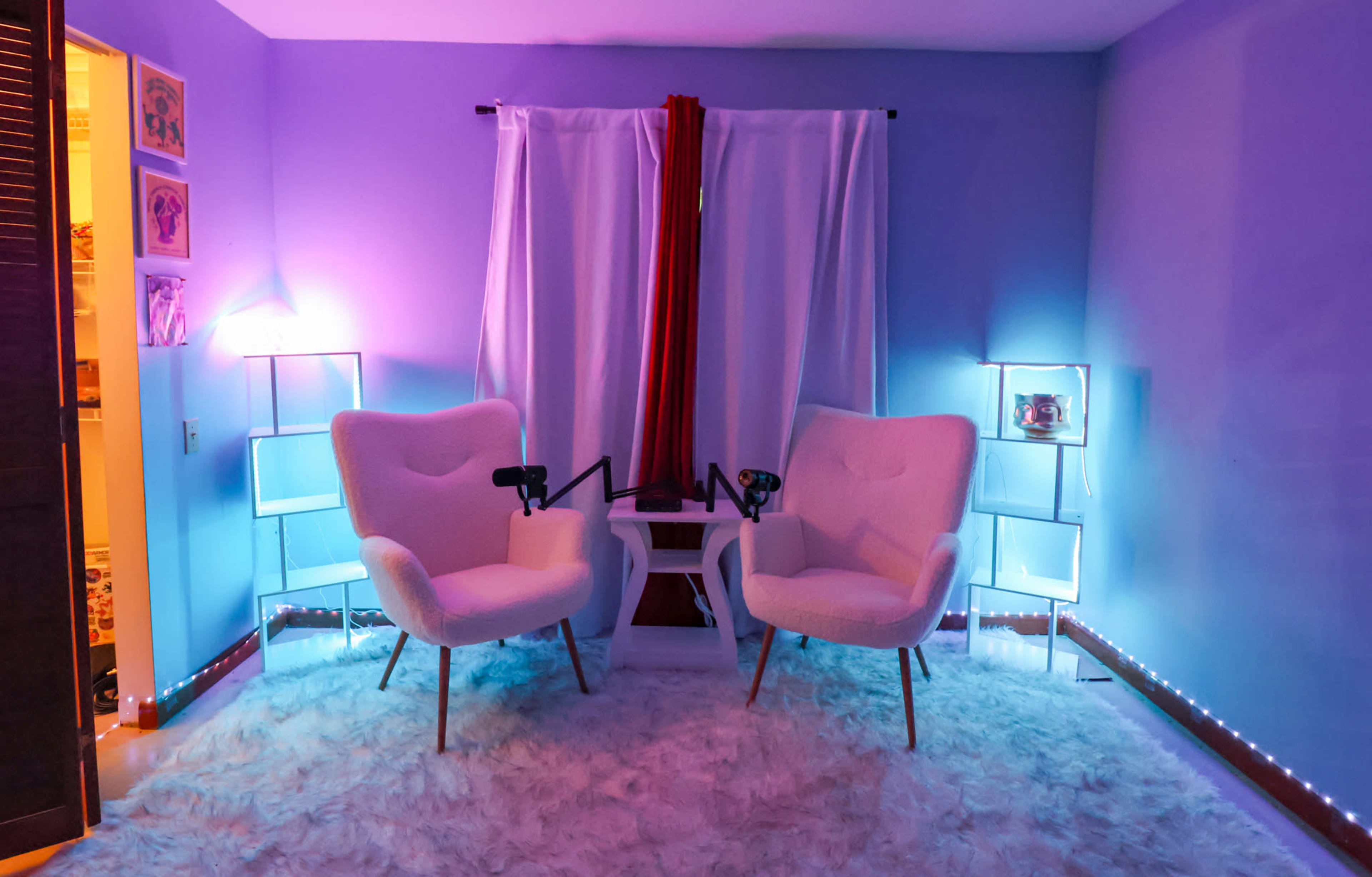 The image shows a brightly lit podcast recording studio with two white chairs facing each other, a small table in between, and soft, illuminated decor against a light purple wall.