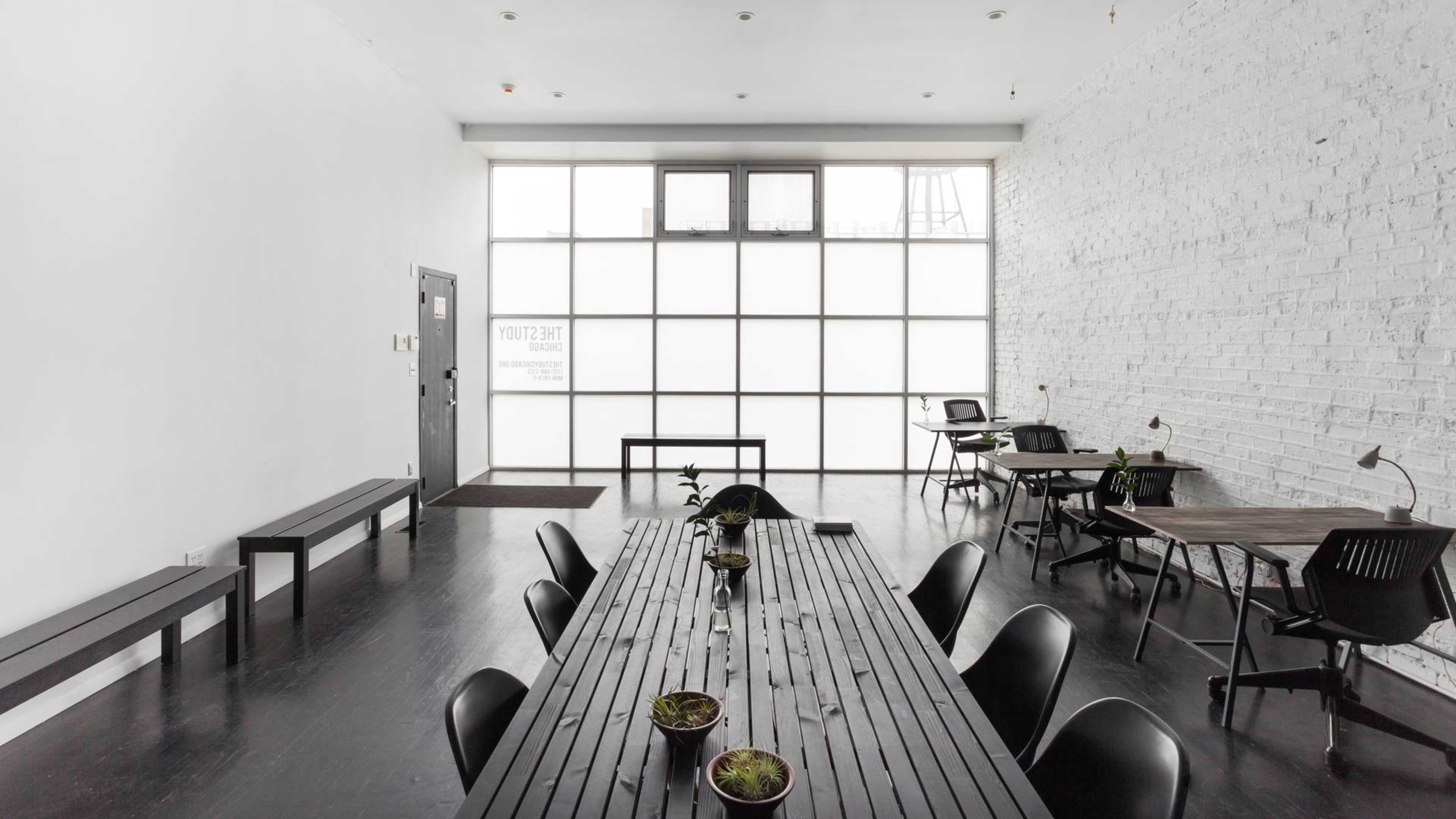 The image shows a minimalist workspace with a long wooden table, several black chairs, and large windows allowing natural light to fill the room.