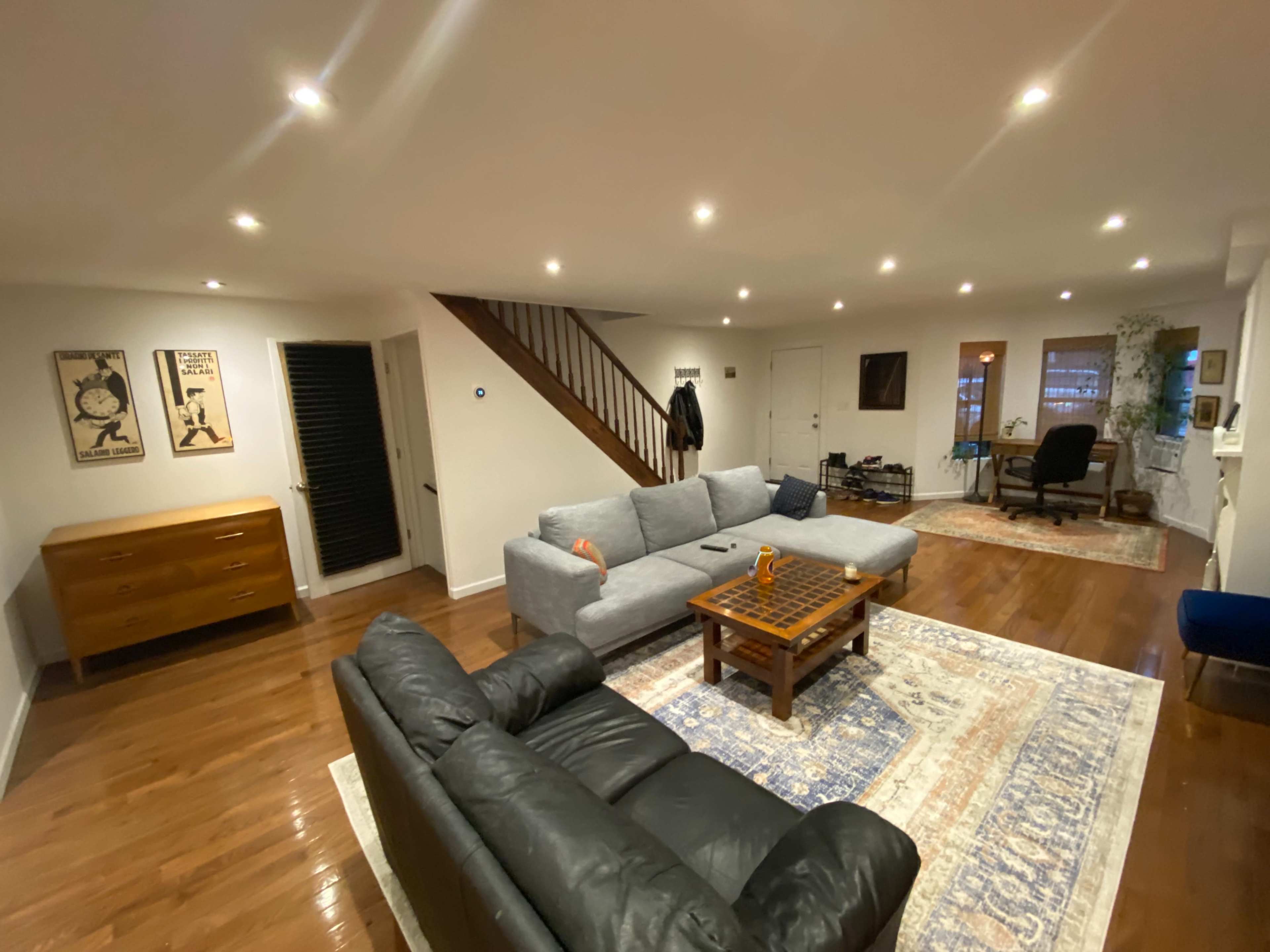 A spacious living room with two couches facing a wooden coffee table, illuminated by recessed lighting, and featuring a staircase in the background.