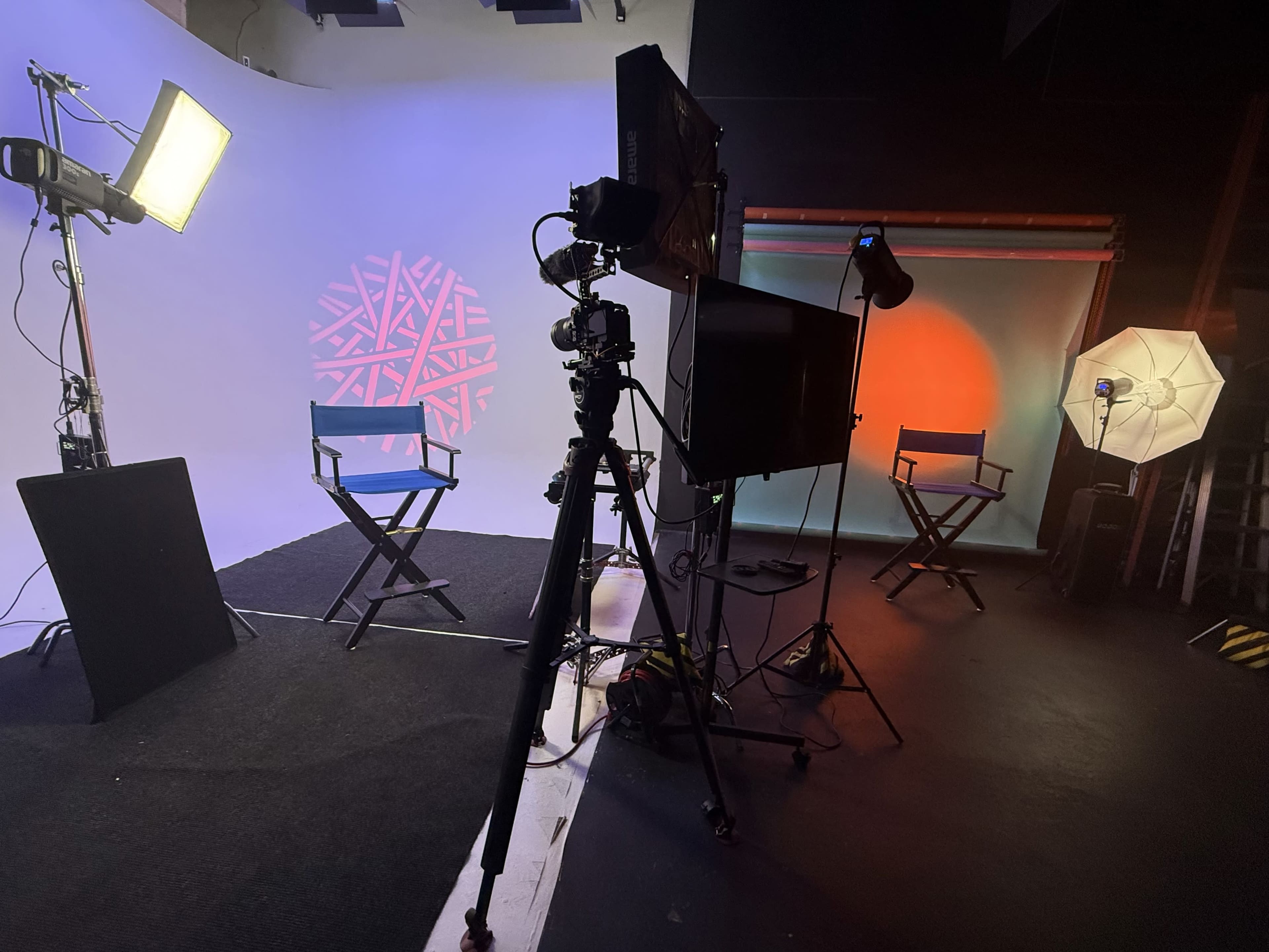 The image shows a photography studio setup with various lights, tripods, and two empty director's chairs against a backdrop featuring colored lighting.