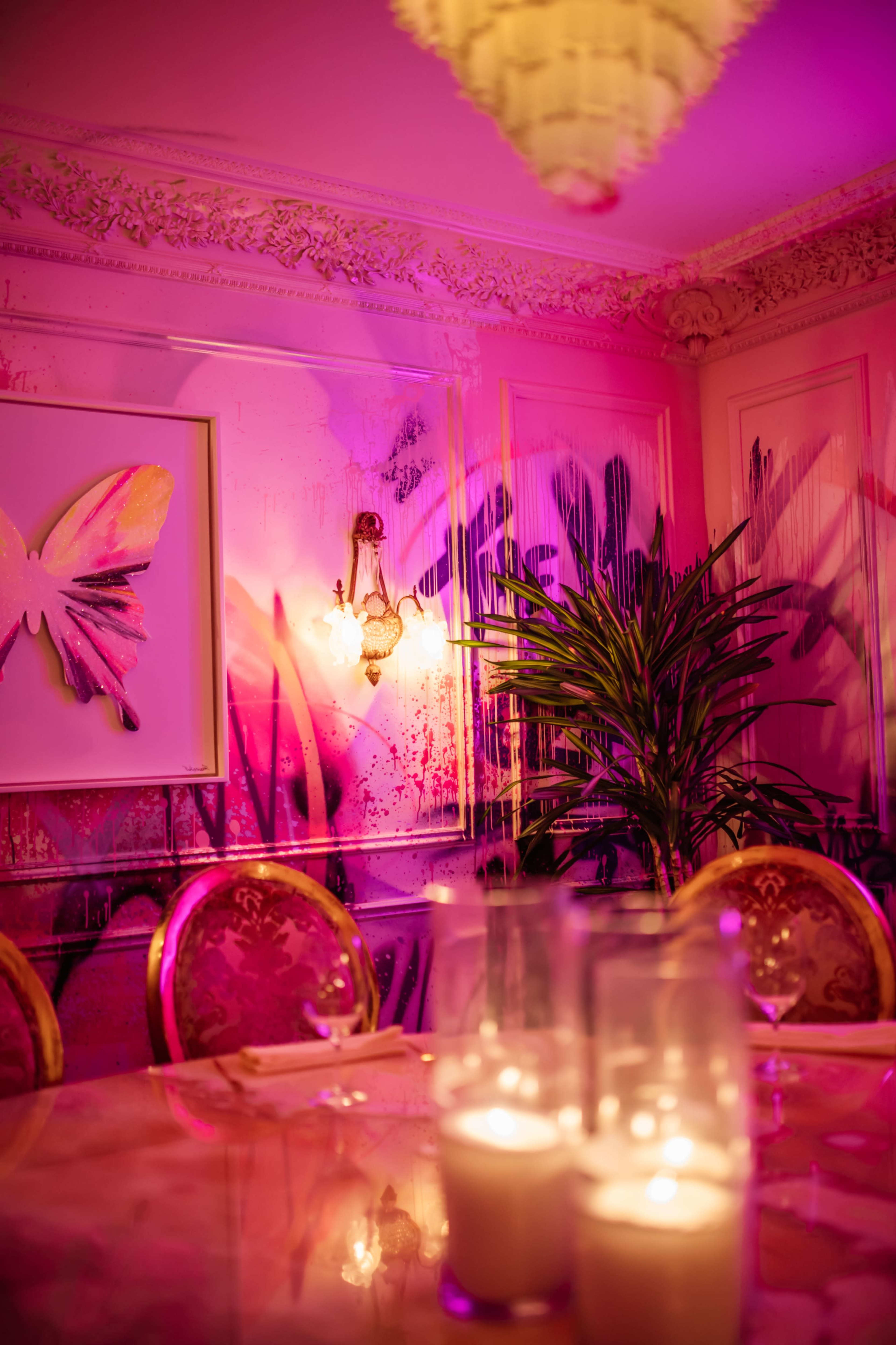 A dining room features a table set with candles, surrounded by upholstered chairs, and illuminated by colorful lighting reflecting off the walls adorned with abstract art and a tropical plant.