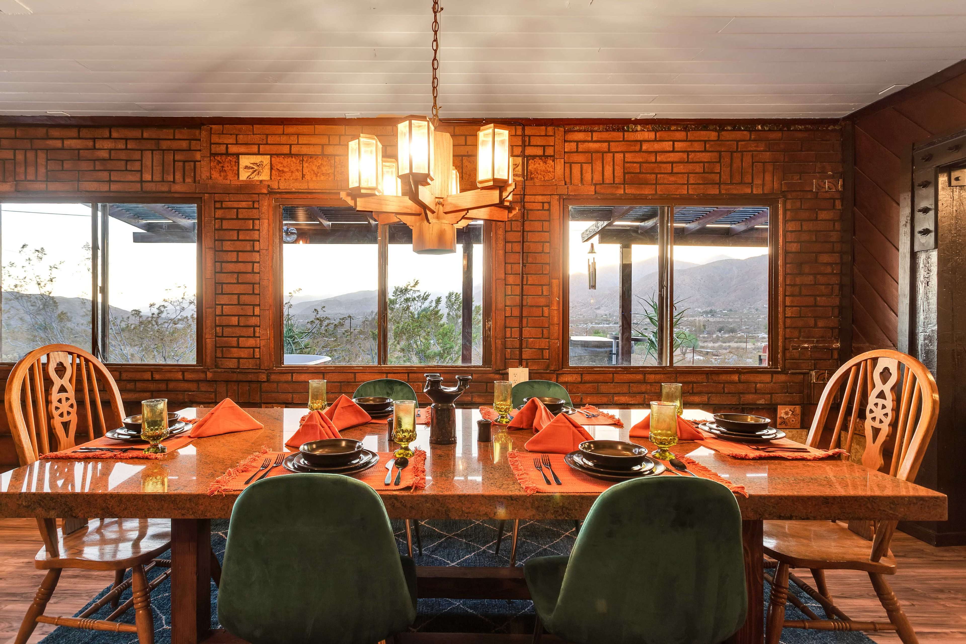 A dining area features a large wooden table set with plates and glasses, surrounded by chairs, against a backdrop of brick walls and windows overlooking a mountainous landscape.