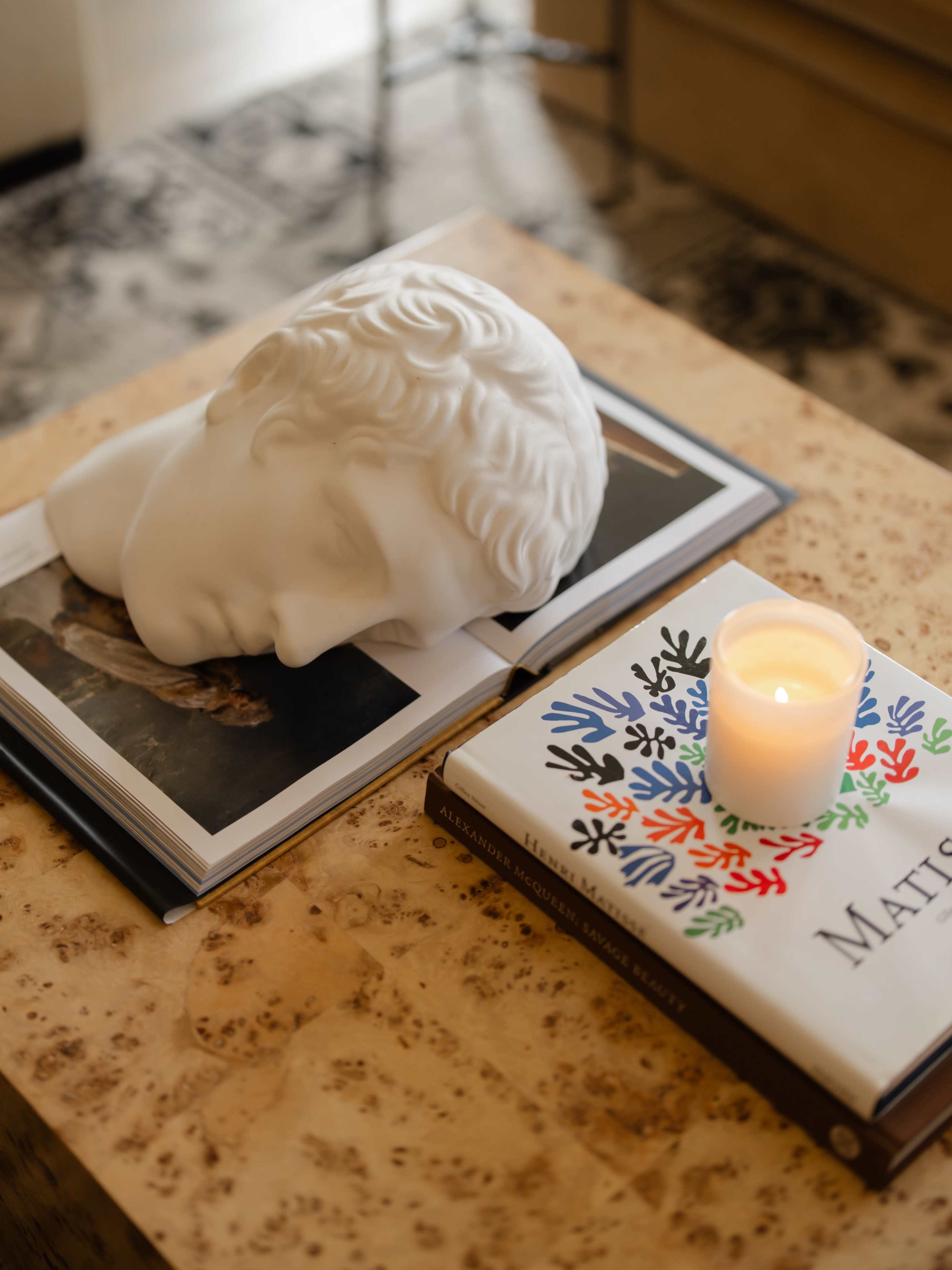 A white sculpture of a head rests on an open book beside another book with a colorful design and a lit candle on a textured, light-colored table.