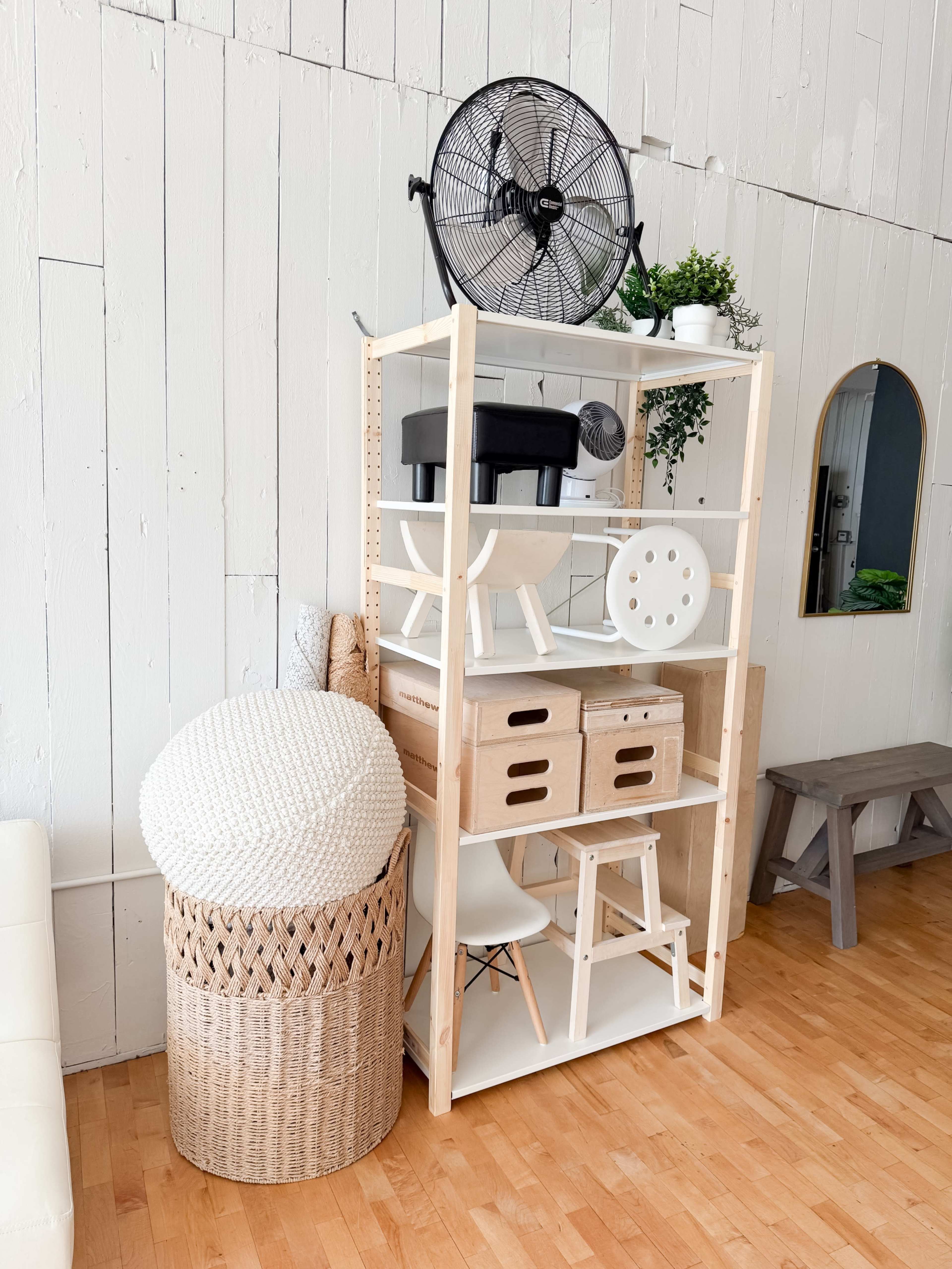 A wooden shelving unit with various items, including a fan, boxes, and decorative plants, is positioned against a white wall in a room with wooden flooring.