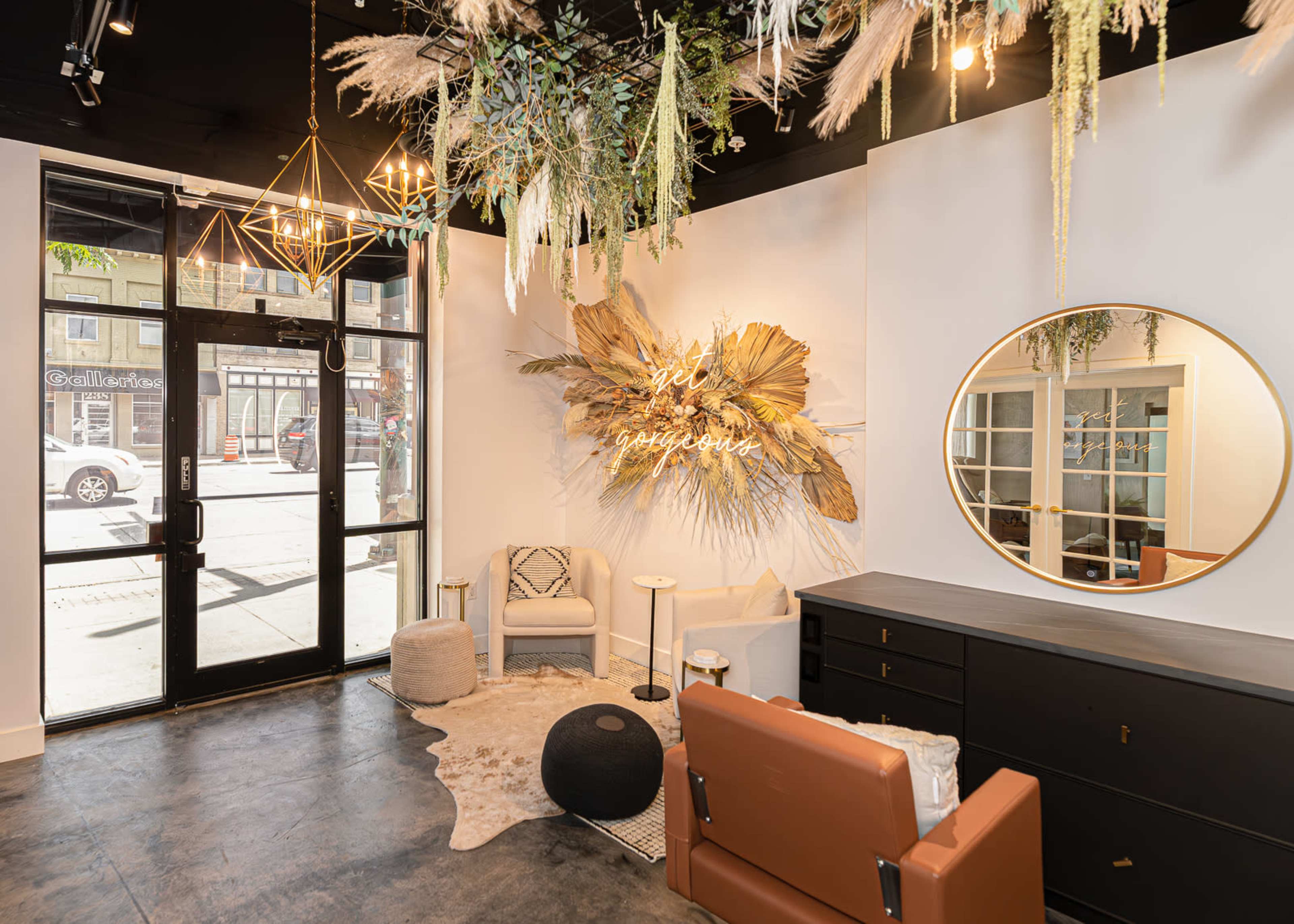 The image shows a modern interior of a salon or boutique featuring a large circular mirror, a decorative wall piece, and greenery hanging from the ceiling, with seating and a black dresser.