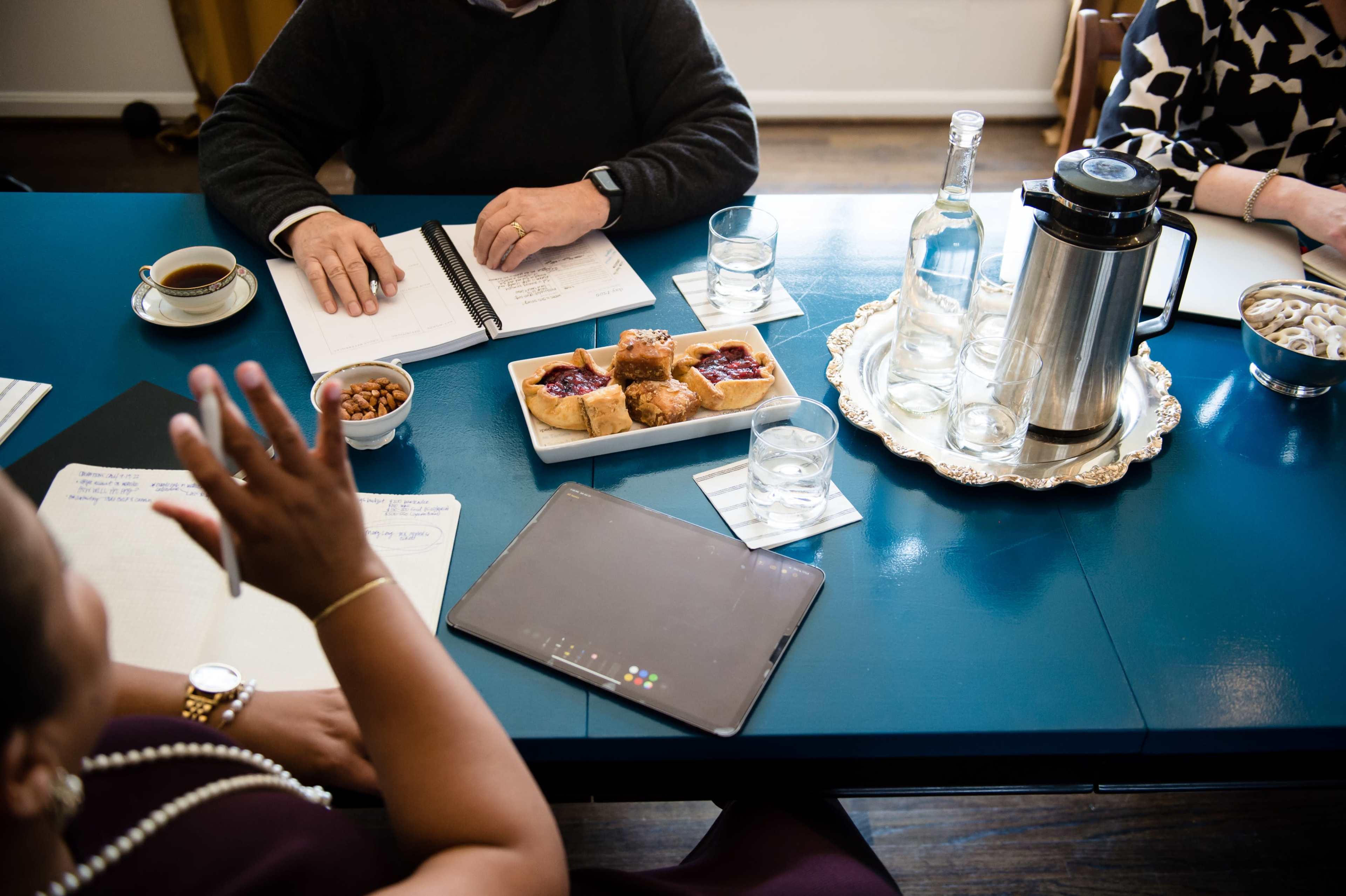 A tabletop setting features a notebook, a tablet, snacks on a platter, drinks, and a coffee pot during a meeting with individuals engaged in discussion.