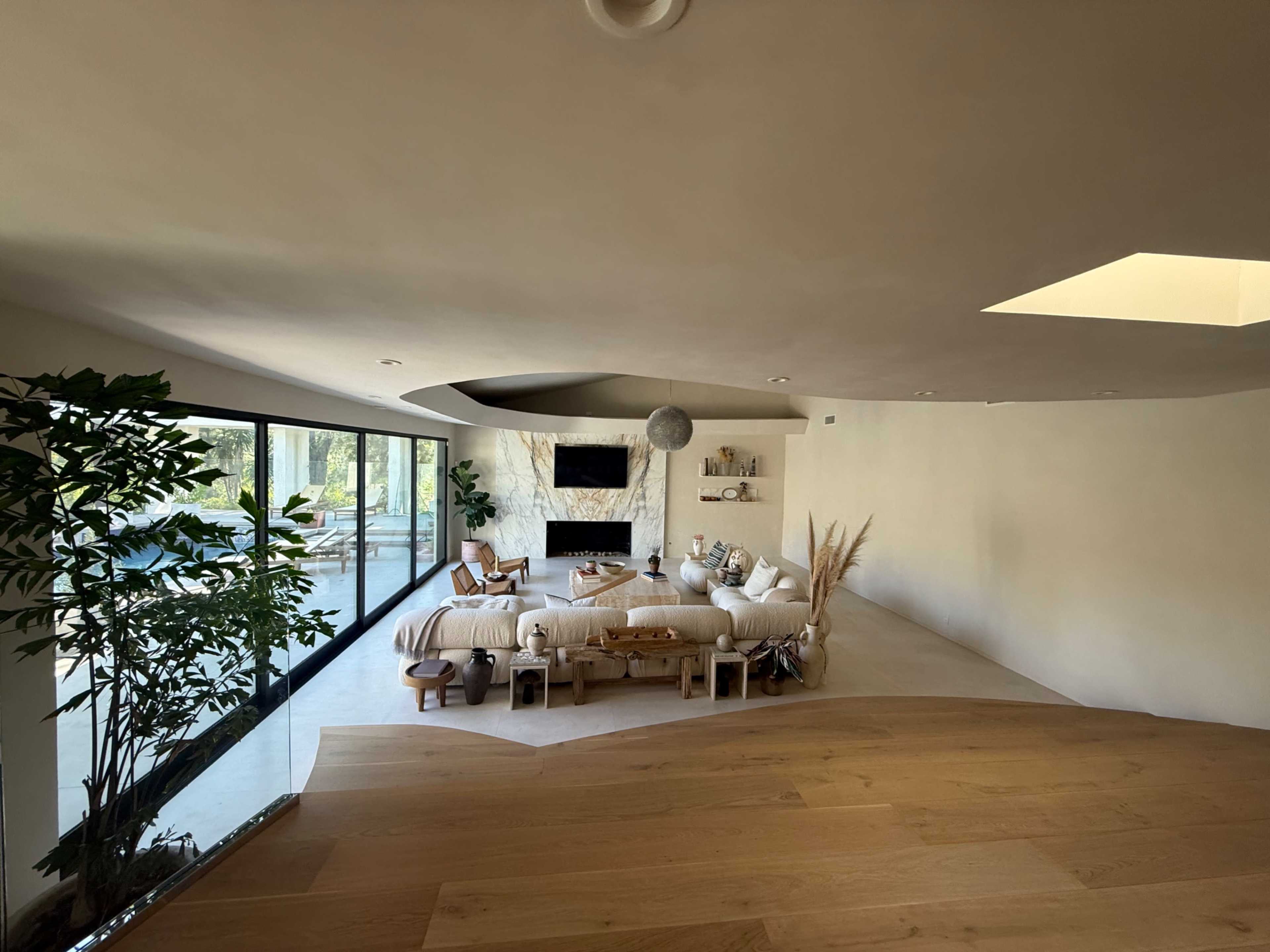 The image shows a modern living room with large glass windows, a plush seating area, and minimalist decor featuring plants and natural materials.