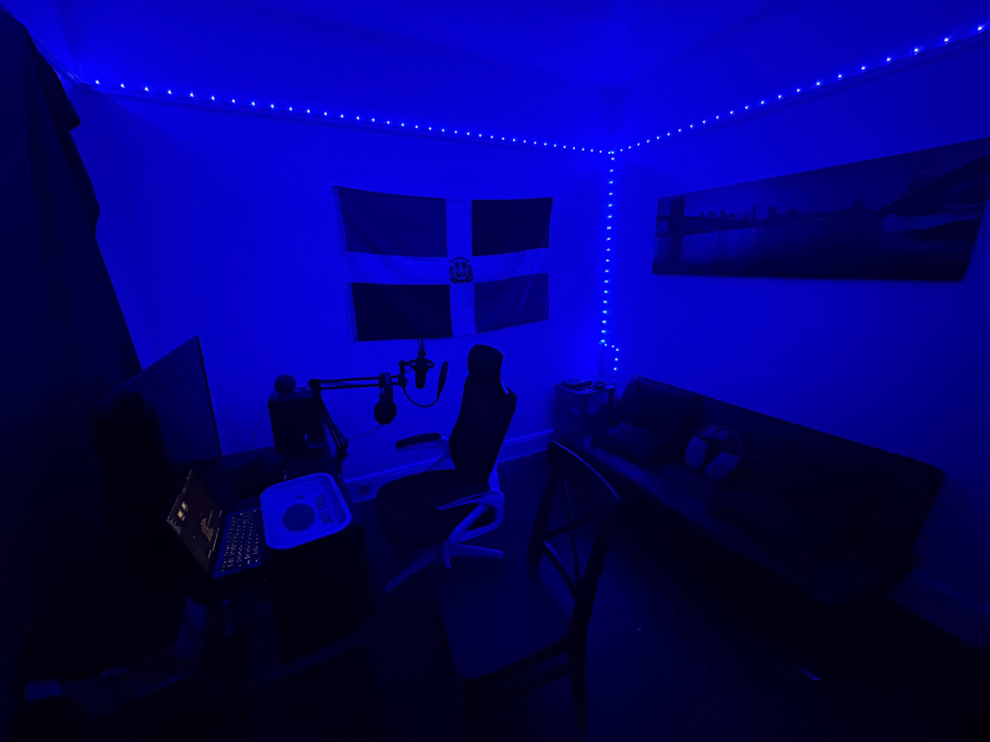 The image shows a dimly lit room featuring a desk with a computer, a black chair, a couch, and a flag hanging on the wall, all accentuated by blue LED strip lighting.