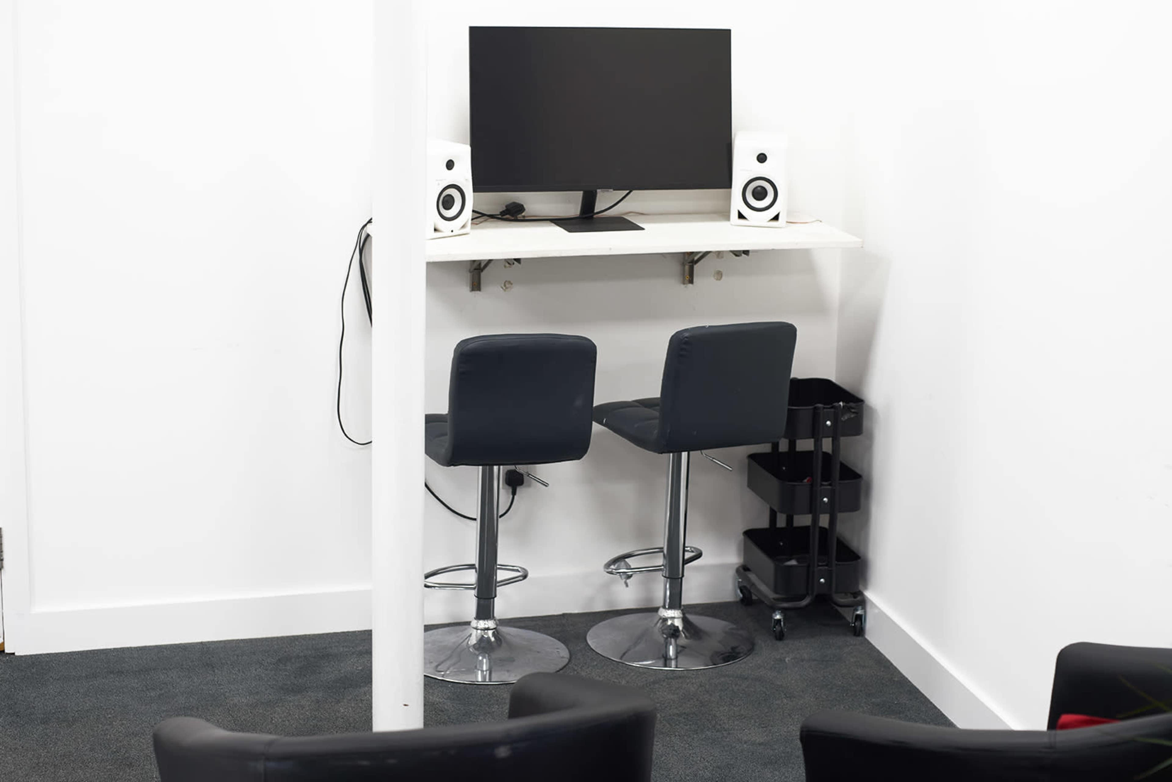 A minimalist office space with a computer monitor mounted on a white wall shelf above two black bar stools and a small rolling cart nearby.