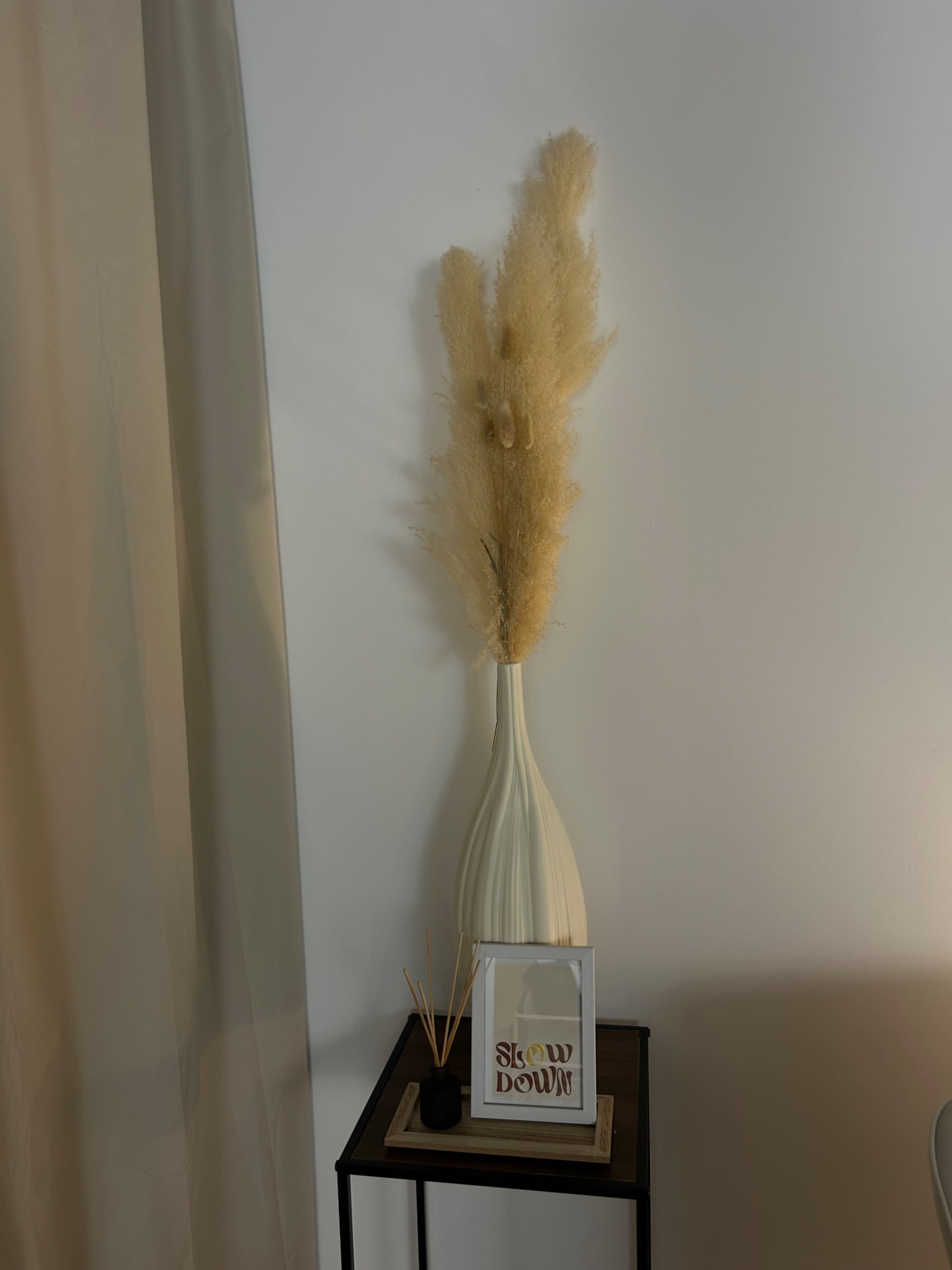 A tall, cream-colored vase holds dried, feathery pampas grass on a small wooden table next to a decorative framed sign that says "Slow Down."