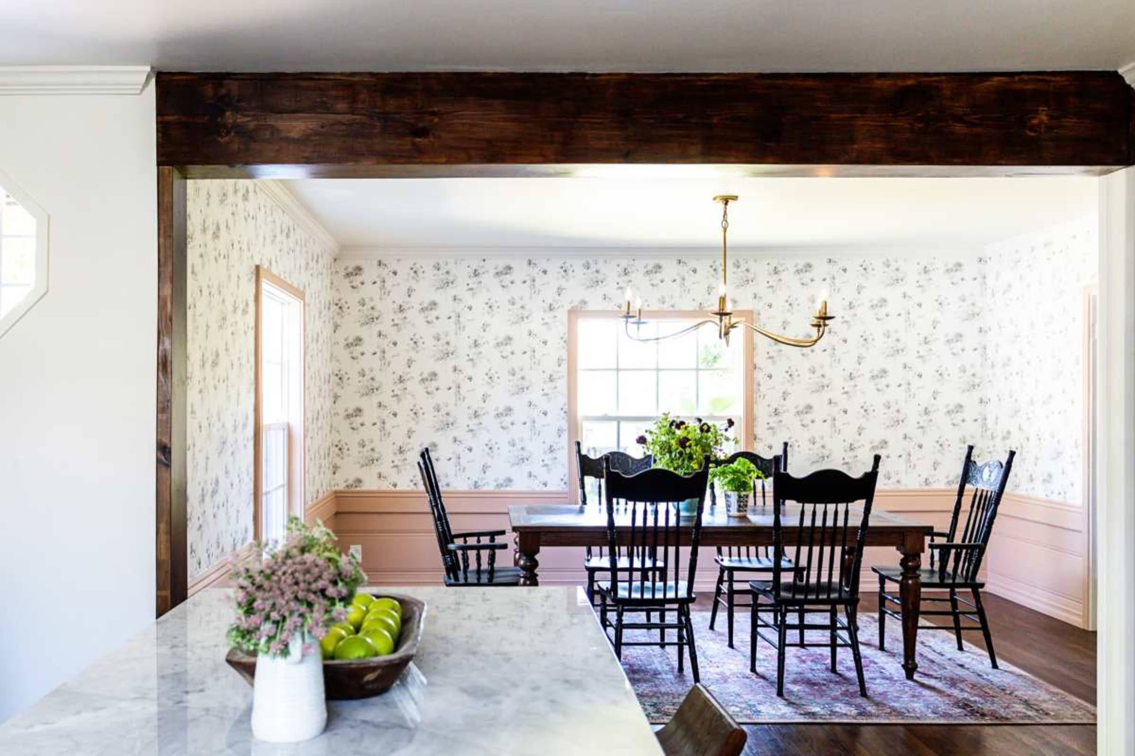 A dining room features a long wooden table surrounded by black chairs, with floral wallpaper and a chandelier above.