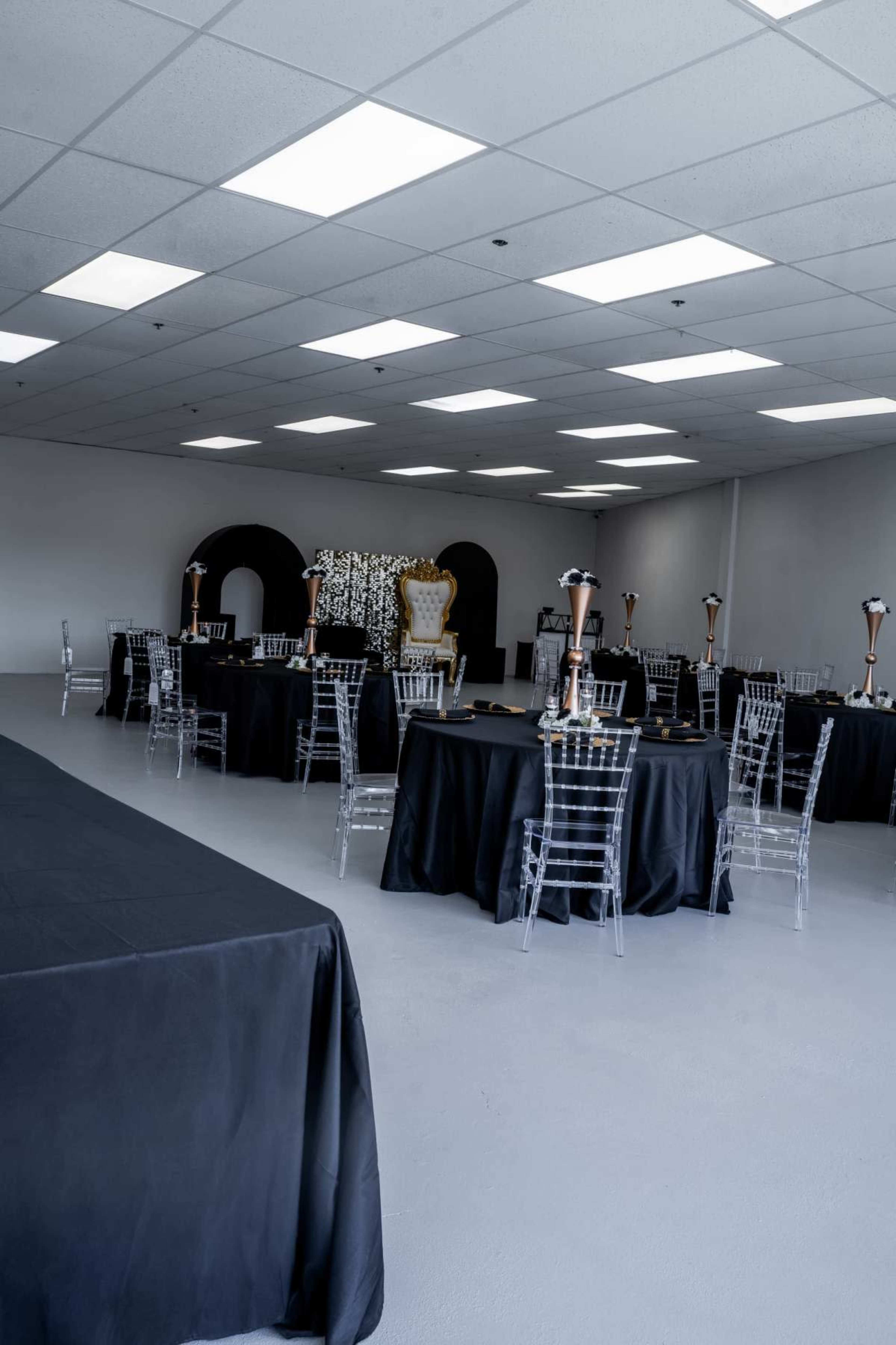 A spacious event room features several round tables set with black tablecloths and transparent chairs, surrounded by modern decor.