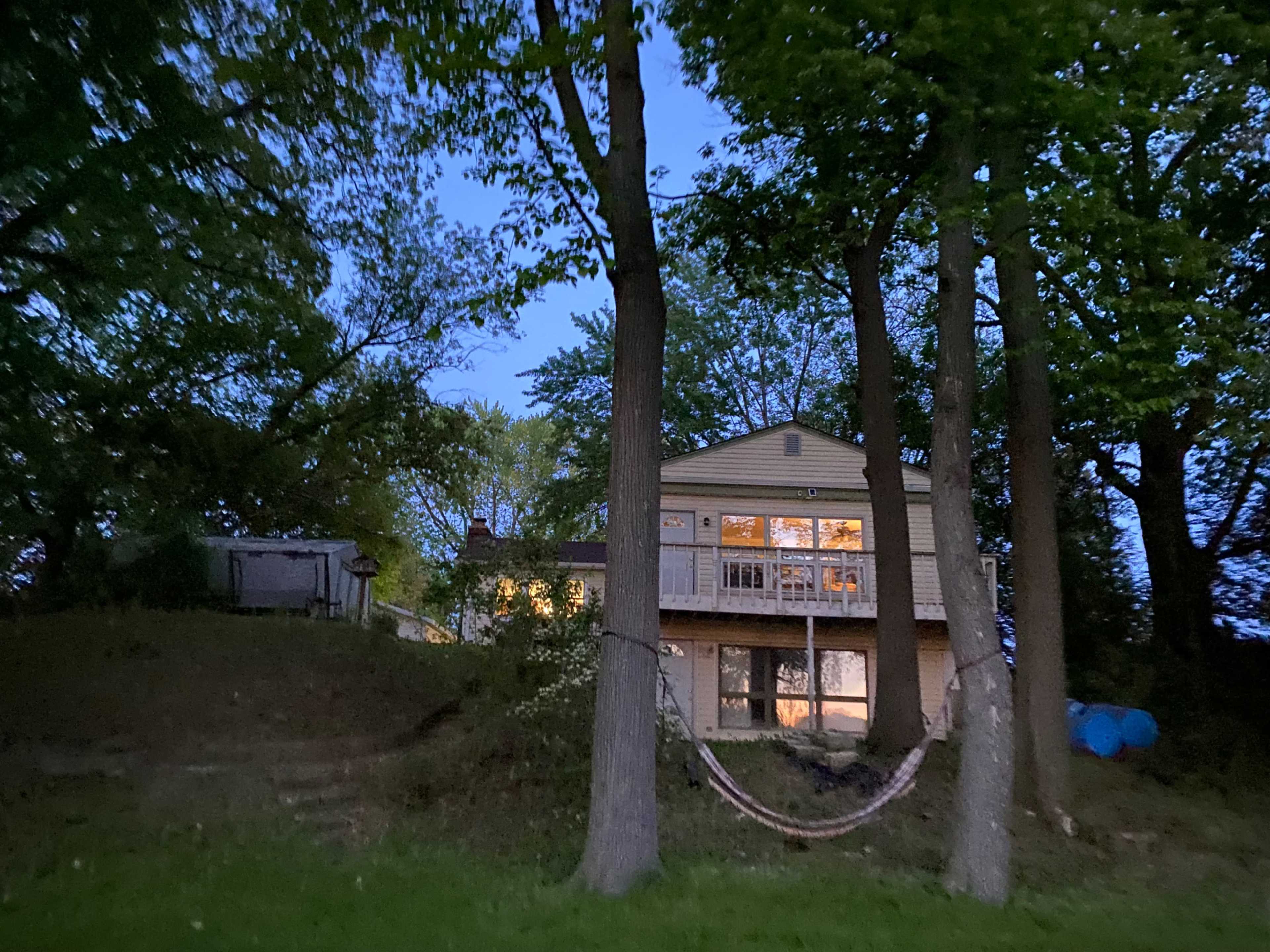 A two-story house is visible among trees at dusk, with a hammock strung between two trunks and warm light coming from the windows.