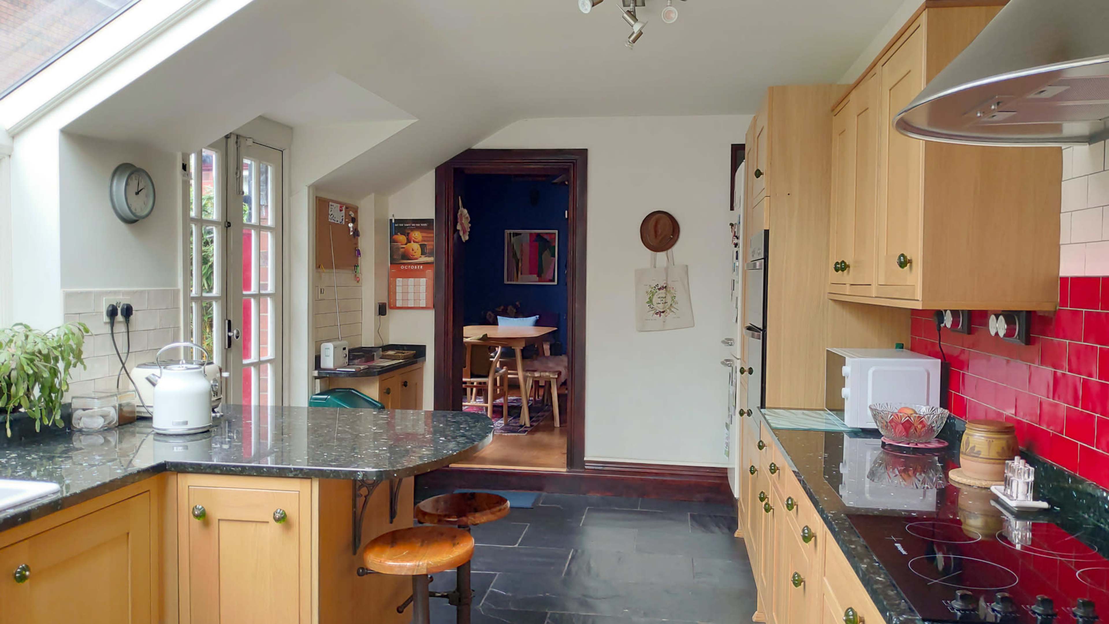 A modern kitchen features wooden cabinets, a black countertop, and red tiled accents, with a door leading to a dining area in the background.