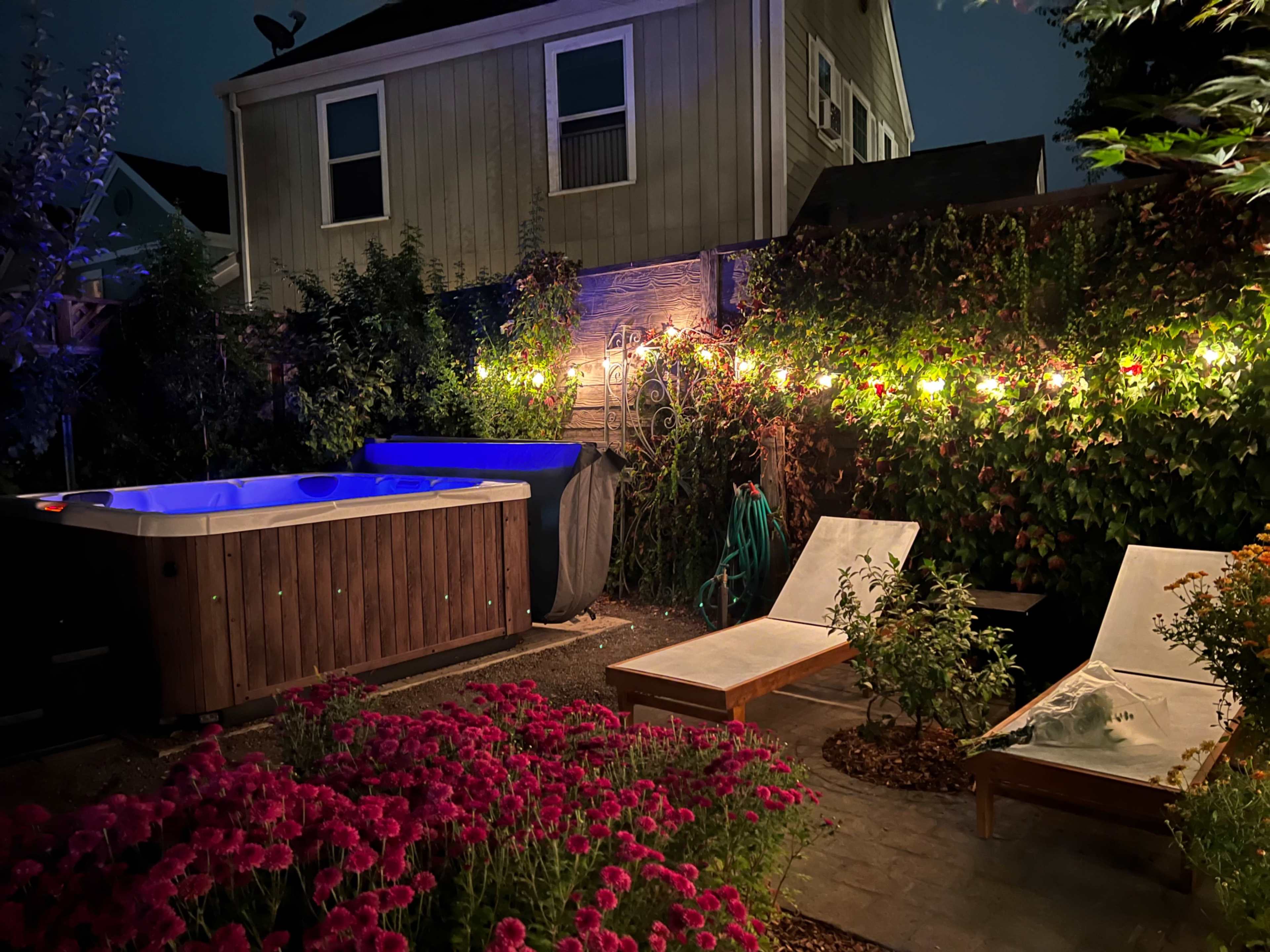 A backyard scene at night features a hot tub, two lounge chairs, and a flower bed with pink flowers, illuminated by string lights along a wooden fence.