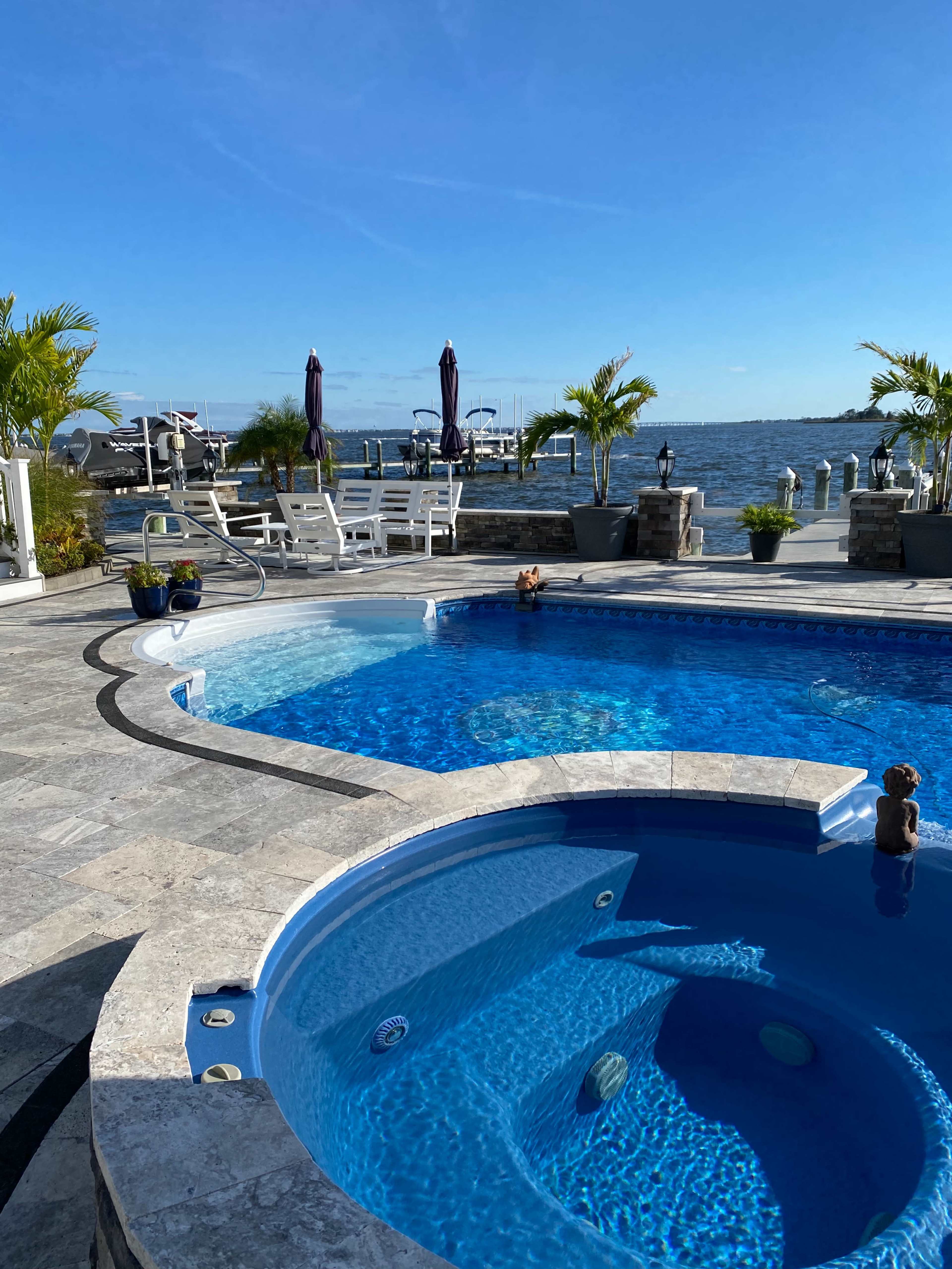 The image shows a swimming pool area with a spa, surrounded by stone paving and overlooking a body of water with boats in the background.