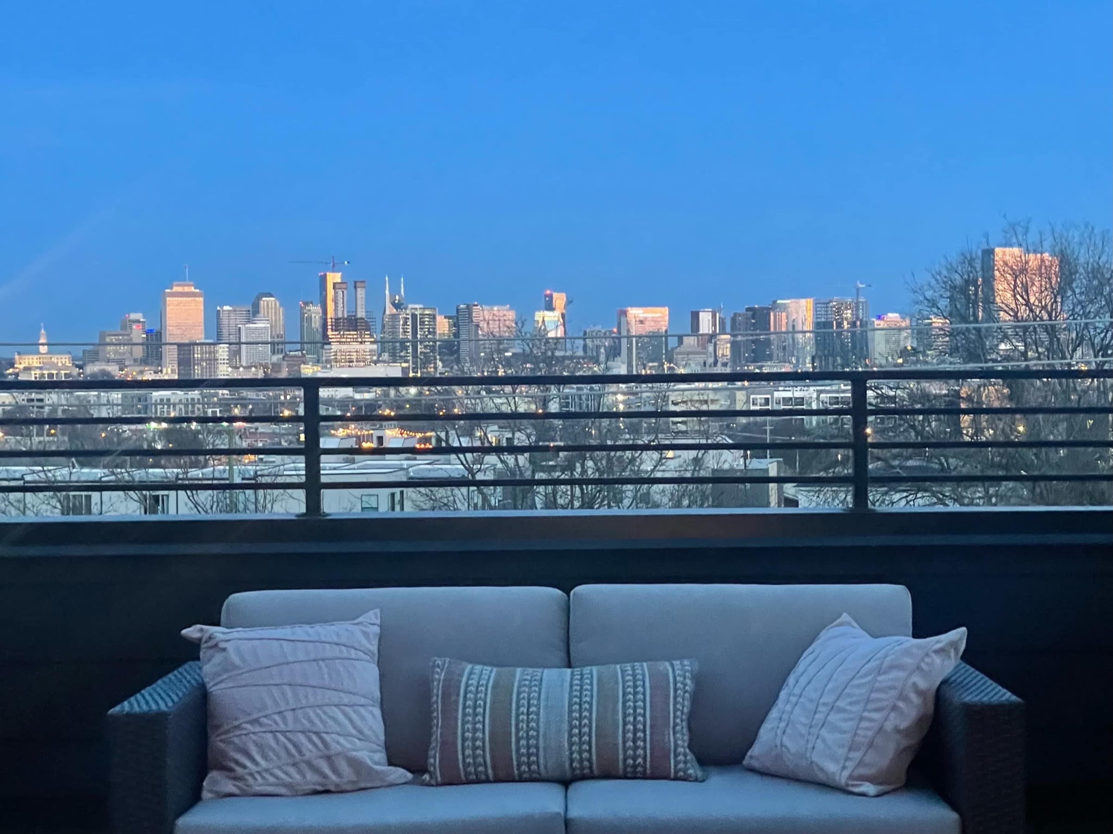 A balcony features a sofa facing a city skyline illuminated at dusk.