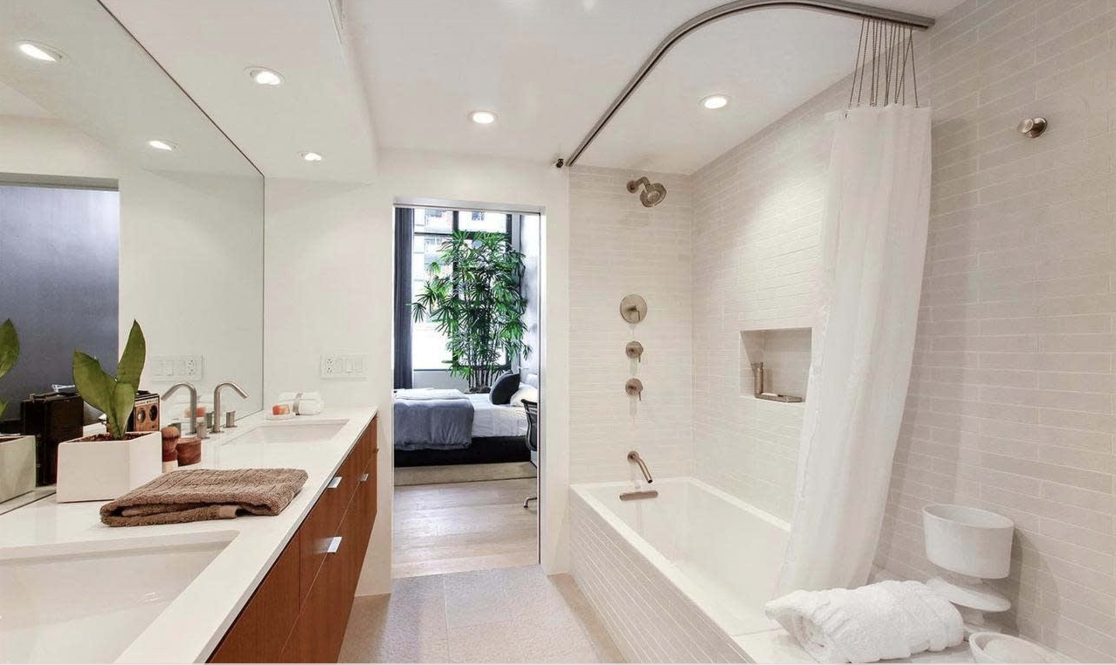A modern bathroom with a bathtub, shower, mirror, and plants visible through a window.