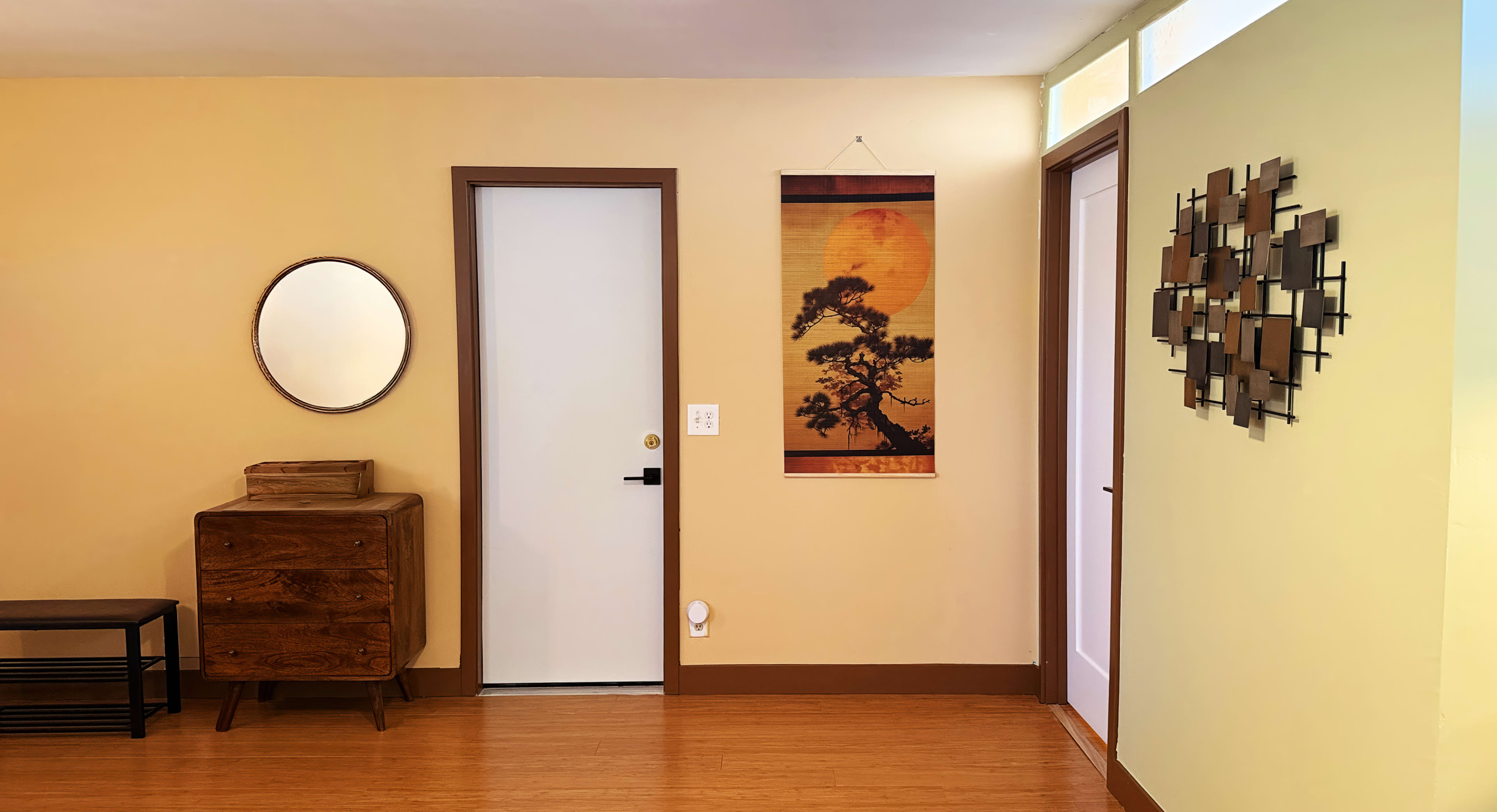 The image depicts a brightly lit hallway with yellow walls, featuring a round mirror, a white door, a wooden chest of drawers, and a wall hanging with a tree and sun design.