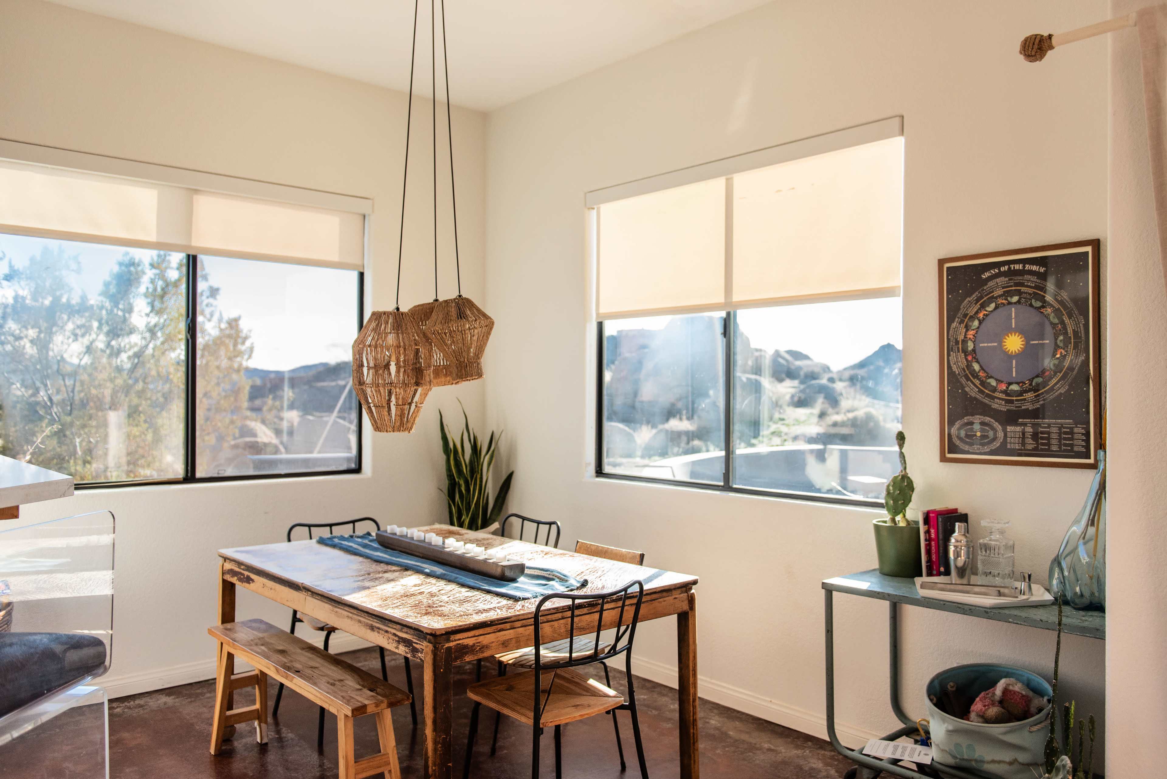 A dining area features a rectangular table surrounded by metal chairs, with large windows illuminating the space and showcasing a natural landscape outside.