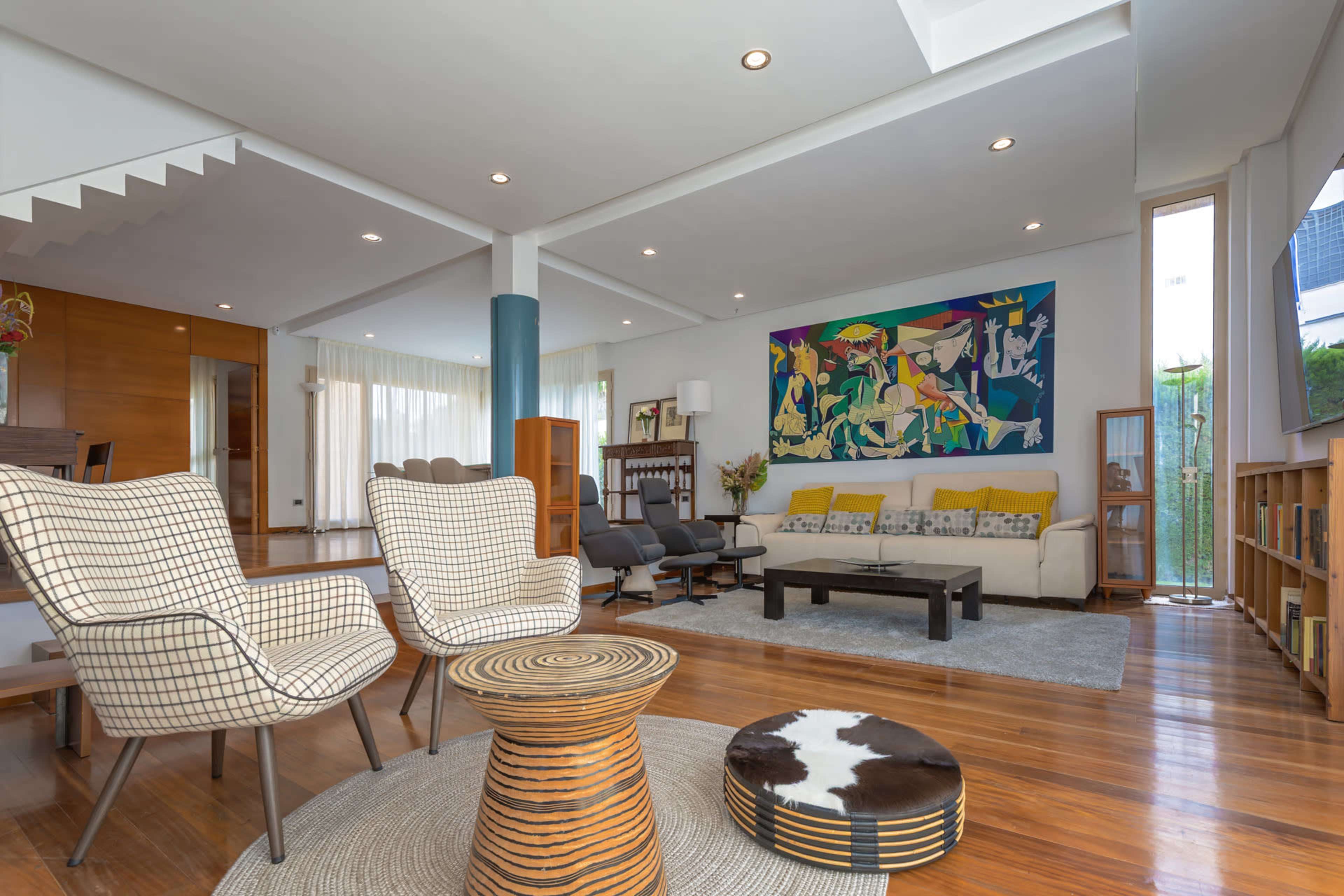 The image shows a spacious living room with wooden flooring, featuring patterned chairs, a low table, a large colorful painting, and large windows allowing natural light.