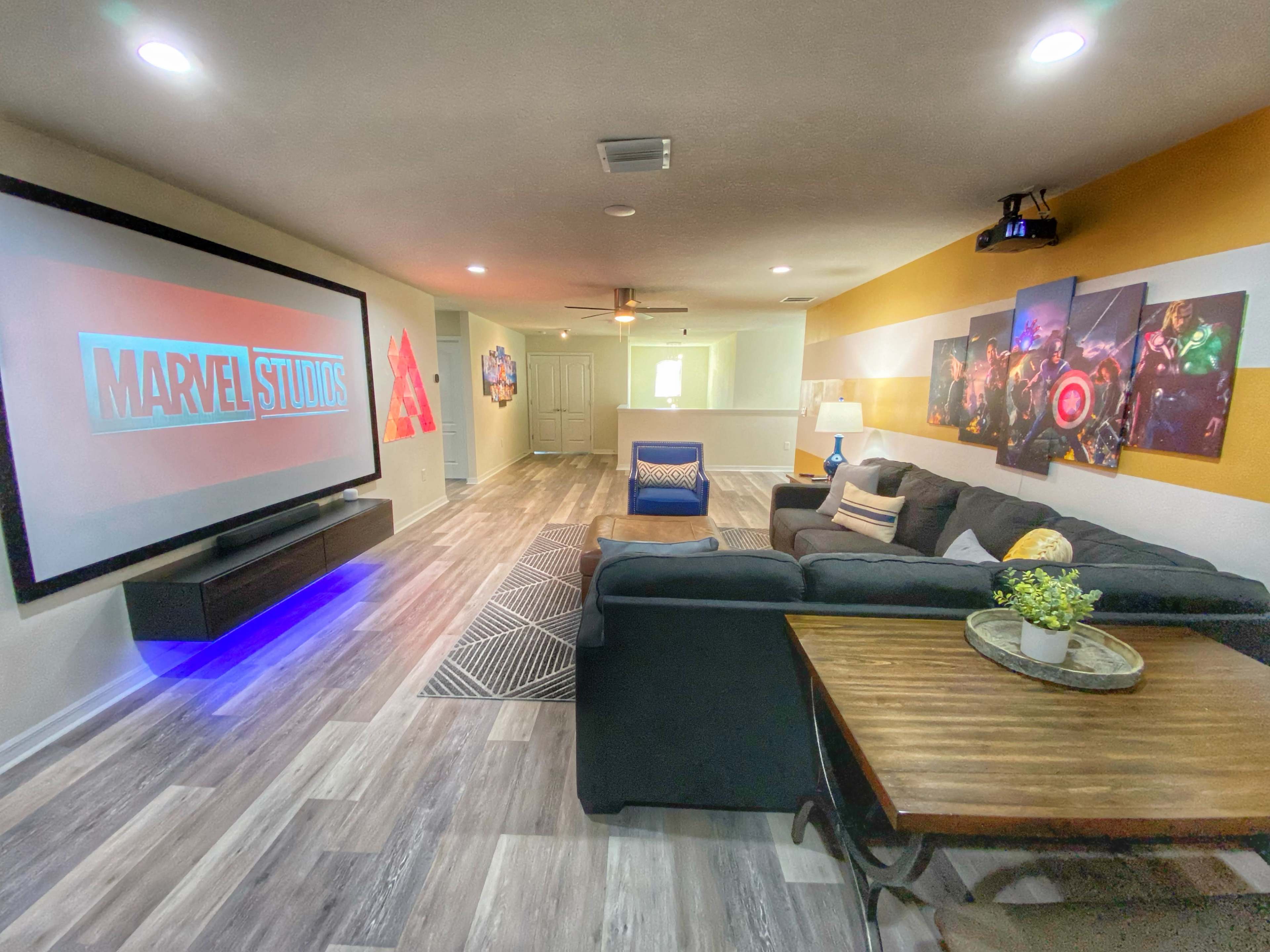 The image shows a modern living room with a large screen displaying "MARVEL STUDIOS," a black sectional sofa, and a coffee table, all set against a backdrop of colorful wall art.