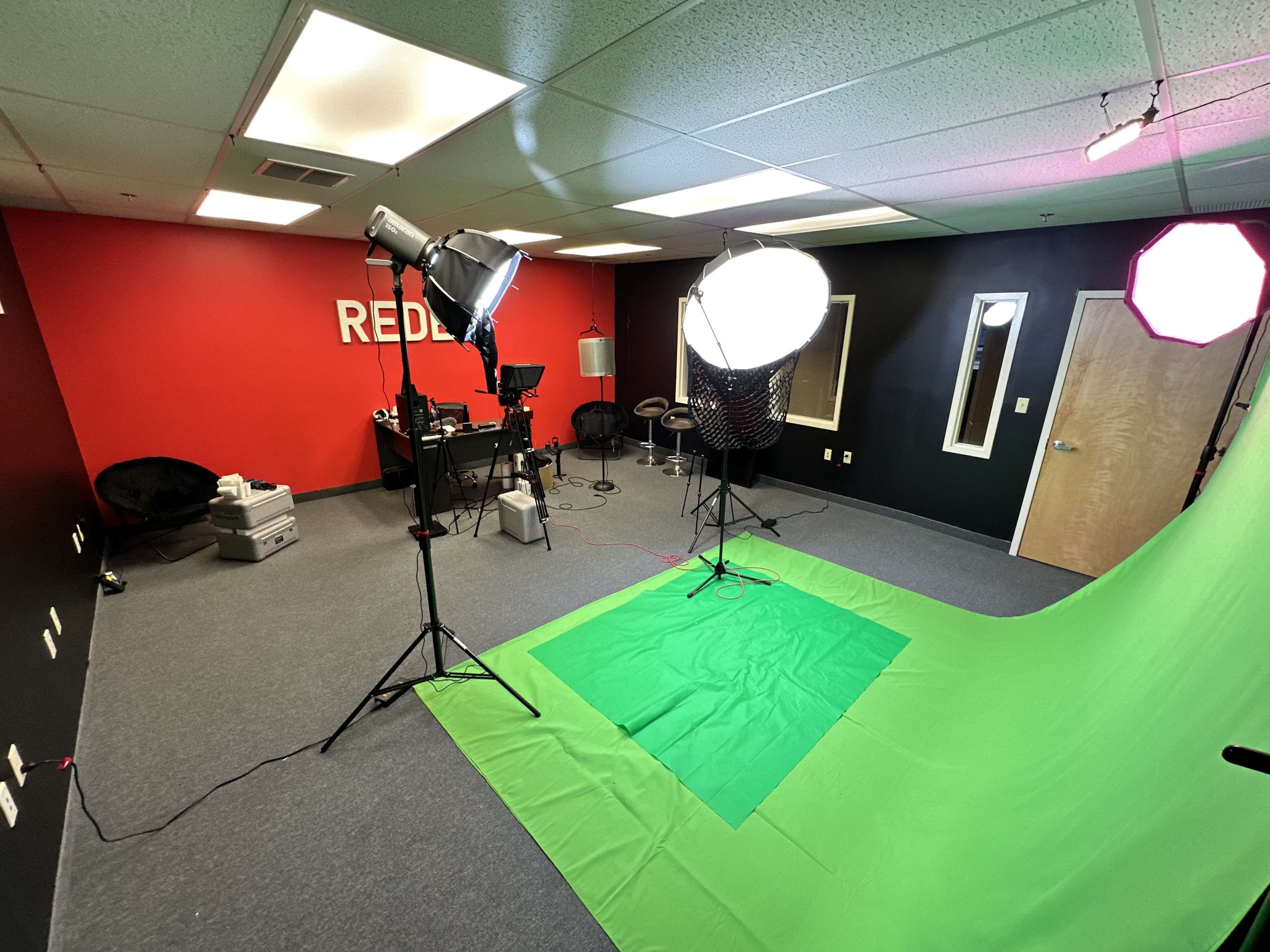 The image shows a studio space equipped with a green backdrop, professional lighting, and various filming equipment against a red and black wall.