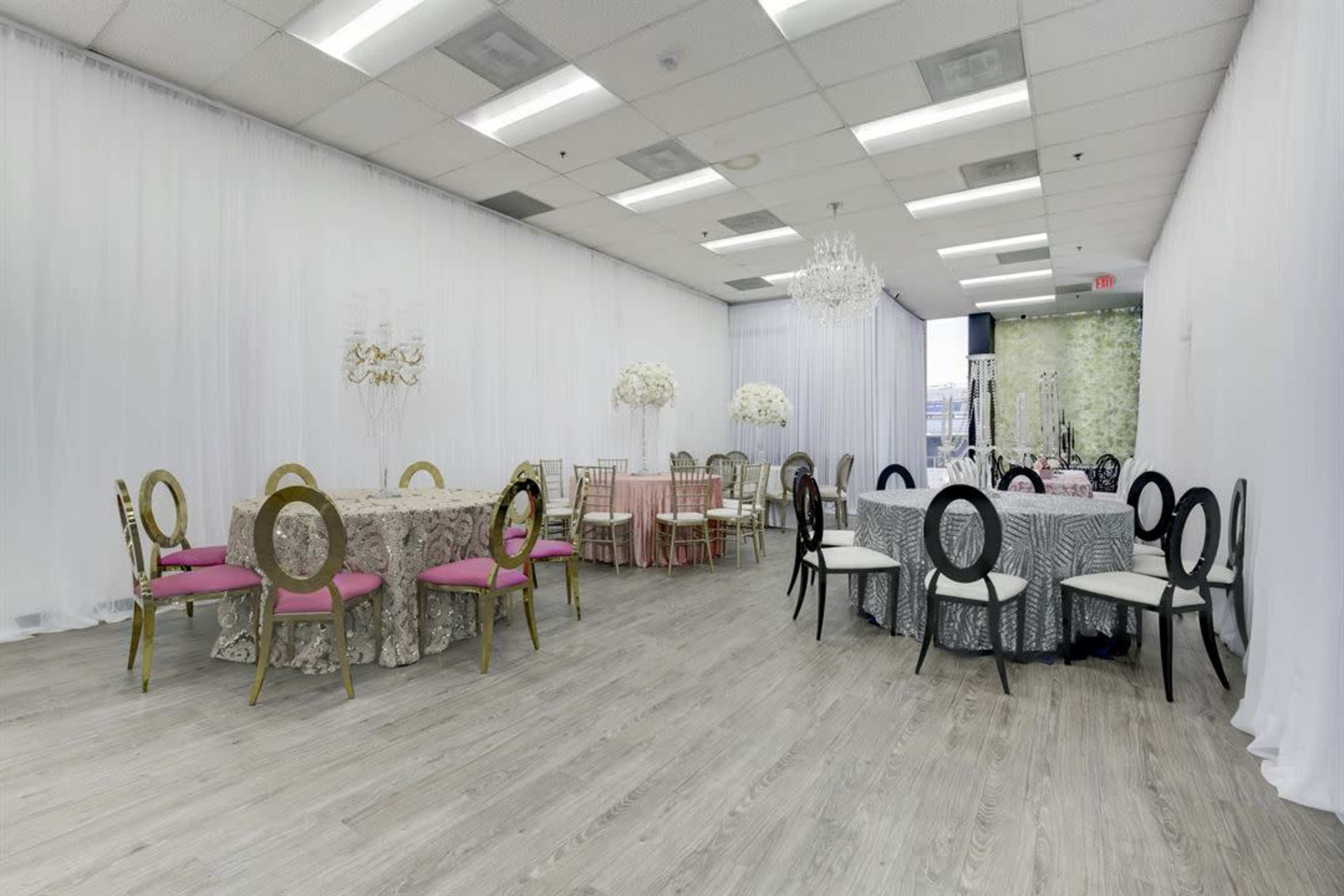 The image shows a spacious event room featuring several round tables covered with elegant tablecloths and surrounded by stylish chairs, with a chandelier overhead and white drapes lining the walls.