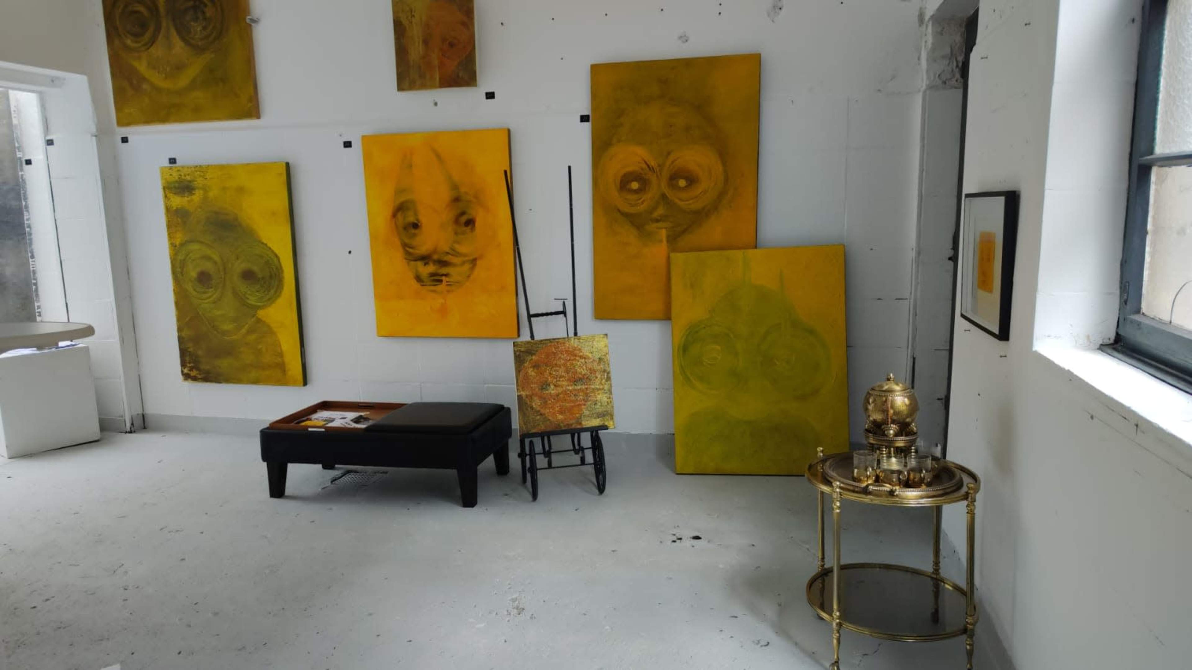 The image shows an art gallery with several yellow abstract paintings displayed on white walls, alongside a small table, a wooden easel, and a gold-colored side table.