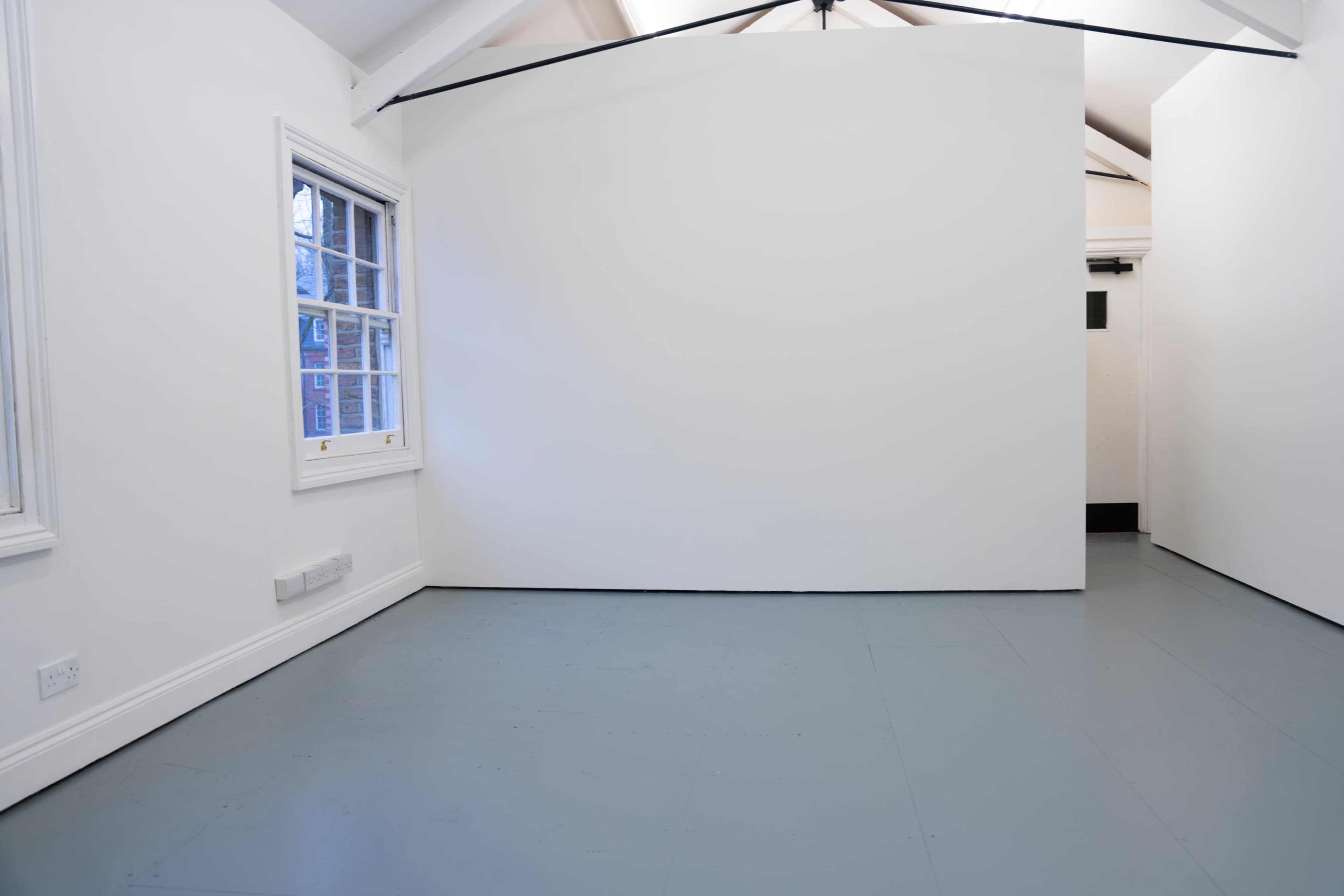 The image shows a minimalistic room with white walls, a gray floor, and a large window on one side.