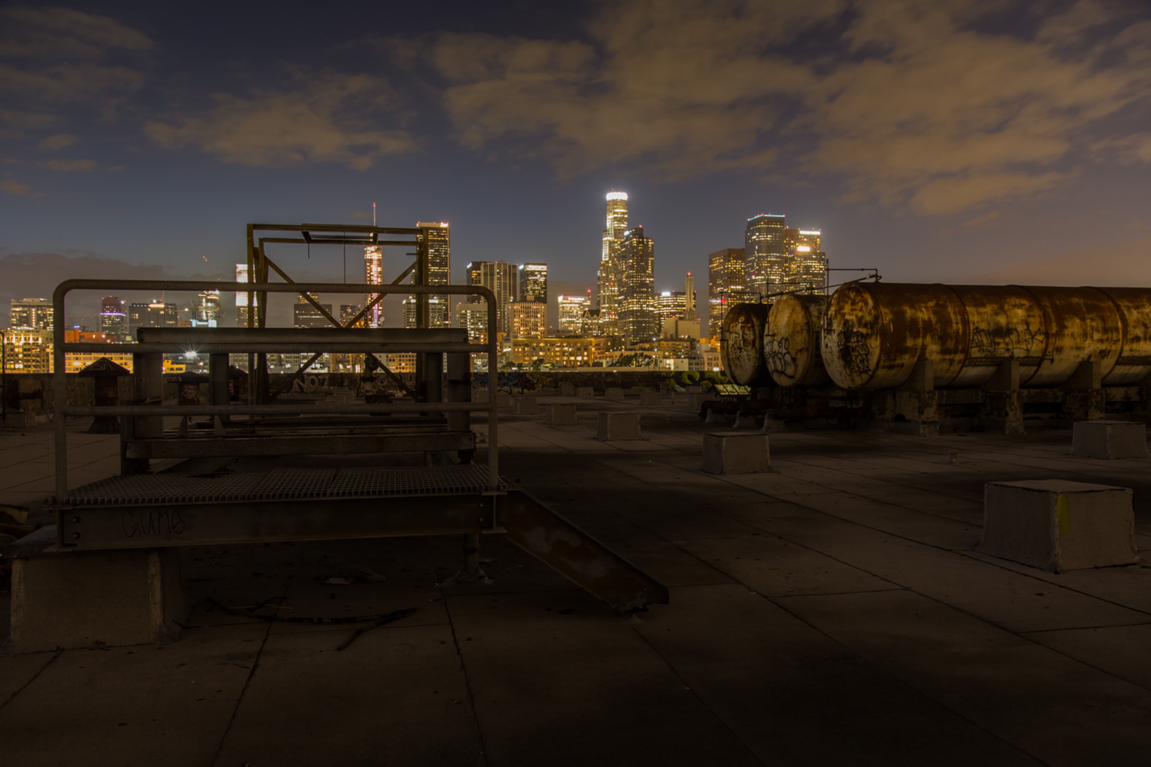 A rooftop view overlooking a city skyline with brightly lit skyscrapers and industrial equipment in the foreground.
