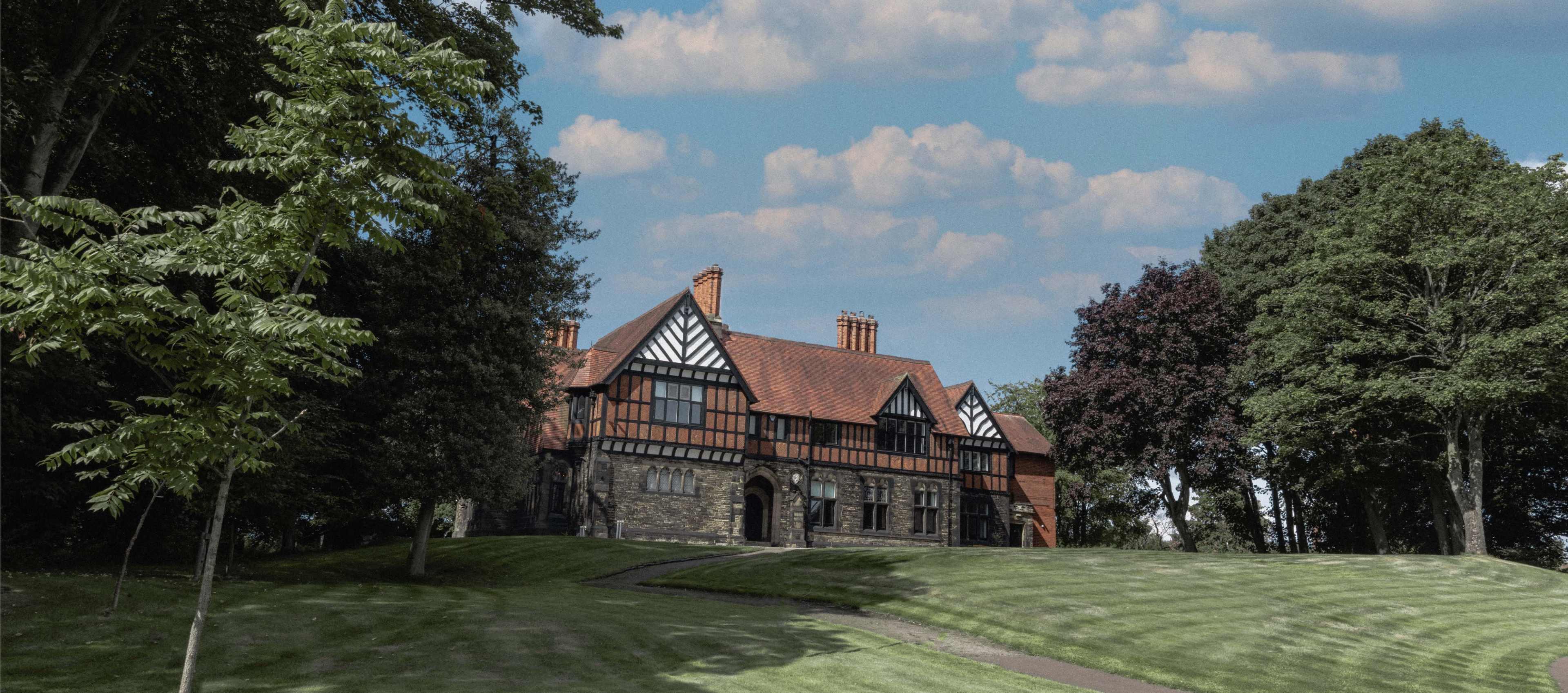 A large, two-story Tudor-style mansion with a red tiled roof is situated on a grassy hill surrounded by trees under a partly cloudy sky.