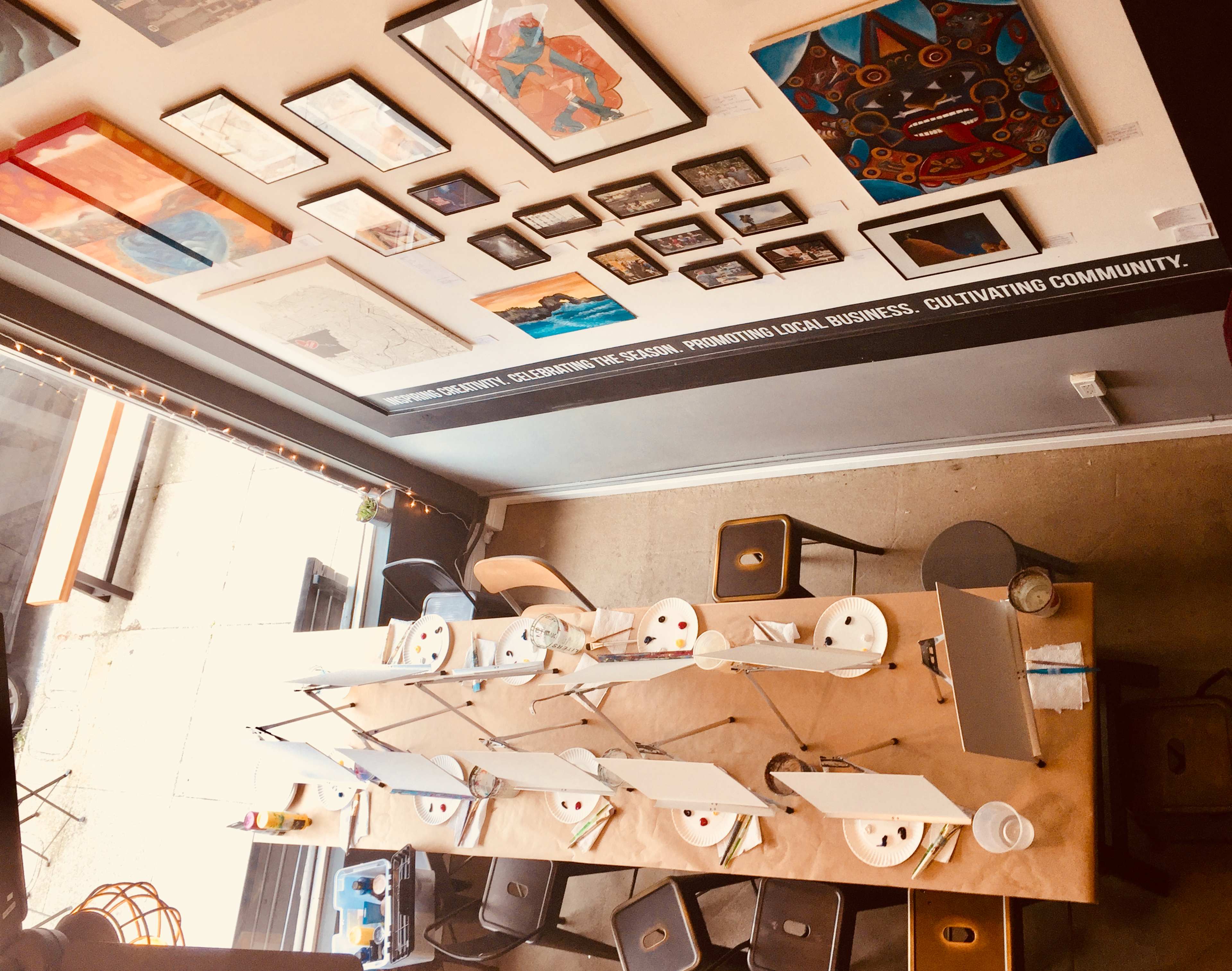 An overhead view of a painting studio featuring a long table set with artist canvases, palettes, and paints, surrounded by chairs and adorned with various framed artworks on the walls.